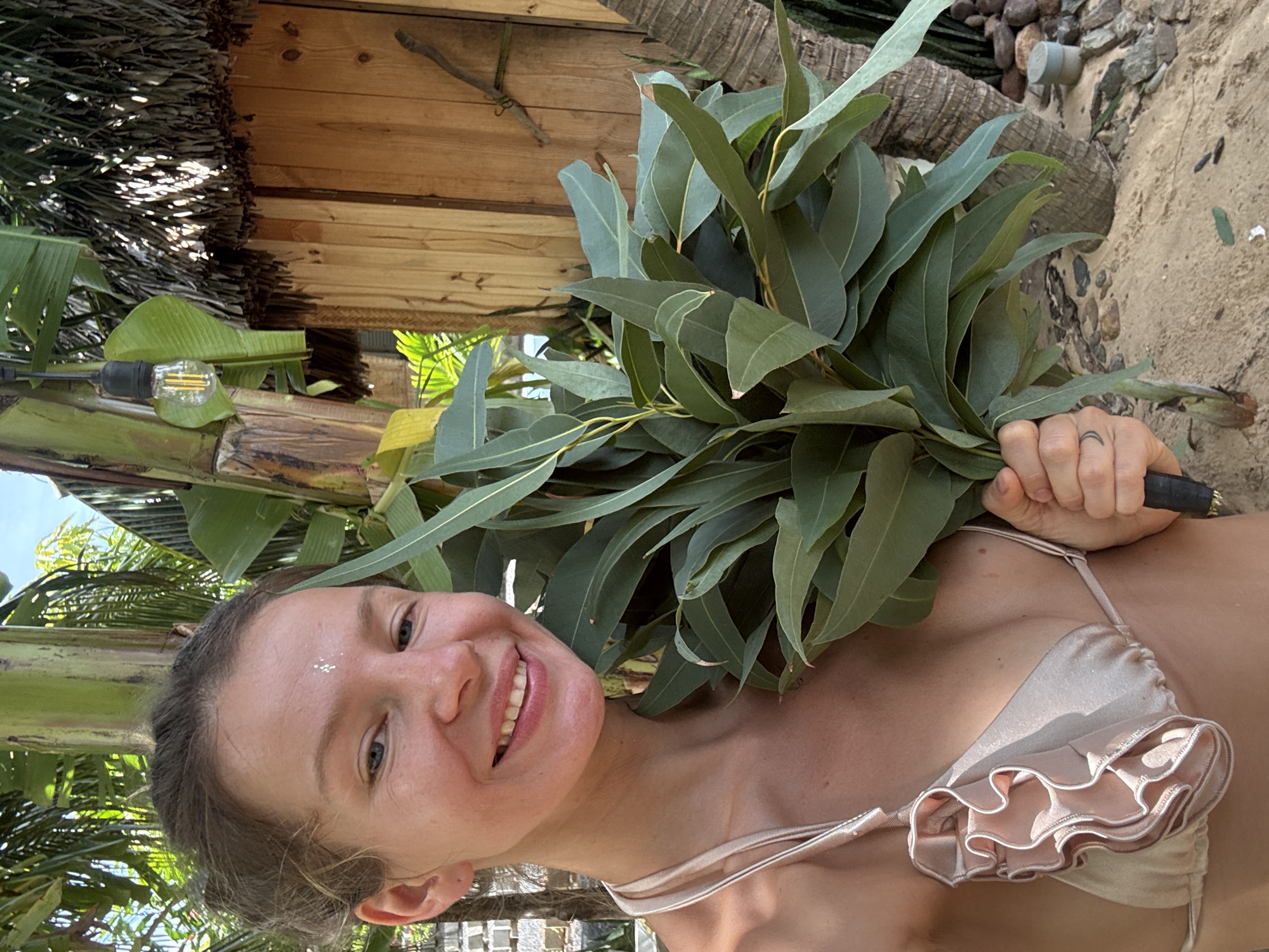 The image shows a woman in a bikini, holding a large bunch of eucalyptus leaves. She is smiling and looking at the camera, with a small glittery decoration on her forehead. The background suggests an outdoor, tropical setting, possibly a beach or resort. There are palm trees and wooden structures visible, as well as sand and rocks. The lighting indicates it is daytime, with bright sunlight casting shadows. The woman's expression and the tropical environment evoke a sense of relaxation and enjoyment. The bikini is light pink or beige with ruffled detailing. Her hand, holding the eucalyptus, shows a ring with a crescent moon design.