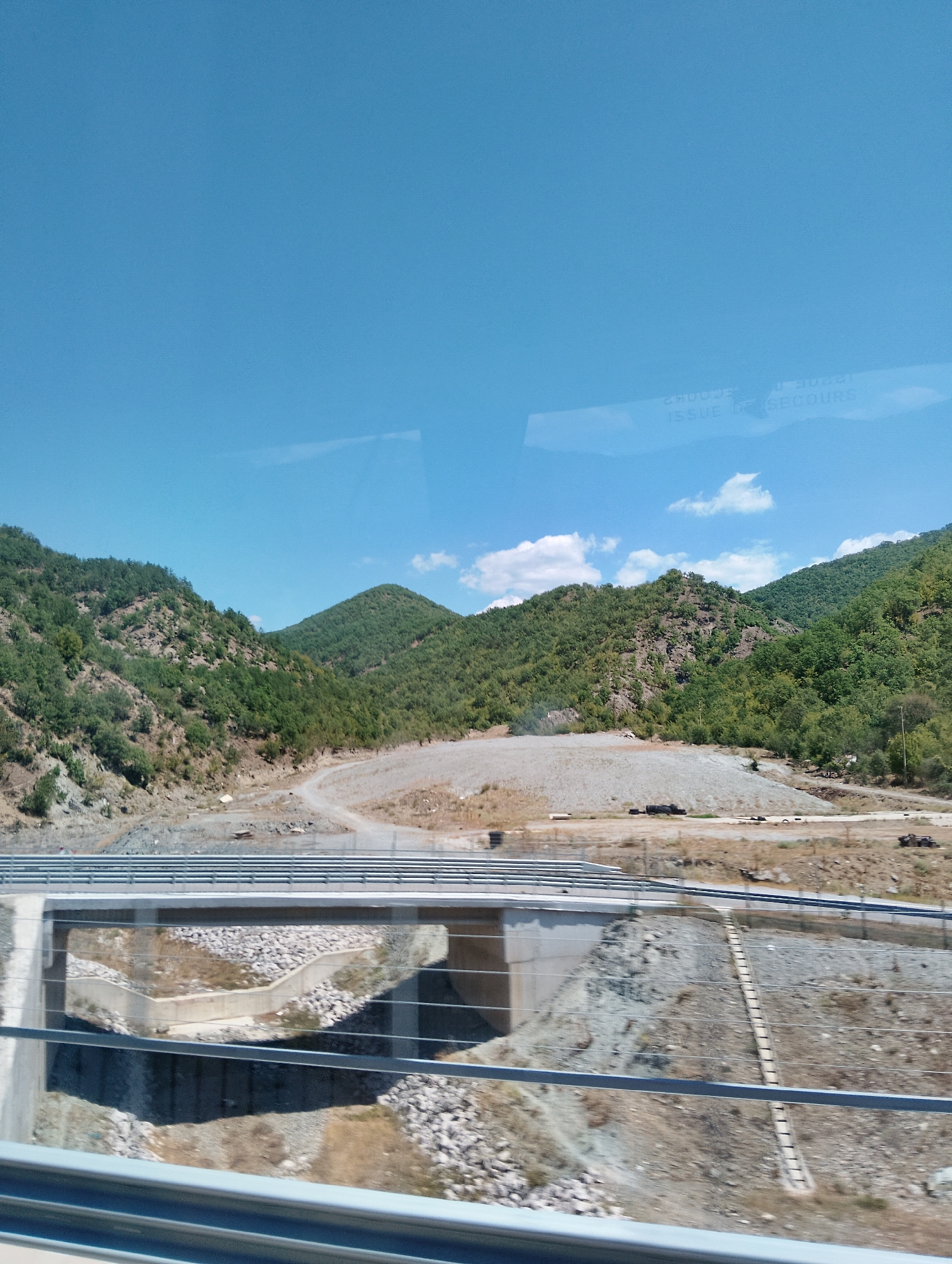 A wide shot captures a scenic view of rolling green hills under a clear blue sky, with a few white clouds scattered across the horizon. In the foreground, a modern concrete bridge spans across a dry ravine filled with rocks. The bridge is flanked by metal guardrails, suggesting it is part of a road or highway. The landscape is generally arid, with patches of dry grass and soil visible around the bridge. The overall scene suggests a rural or mountainous area. The image appears to be taken from a moving vehicle, as indicated by the slight blur and reflections on the glass.