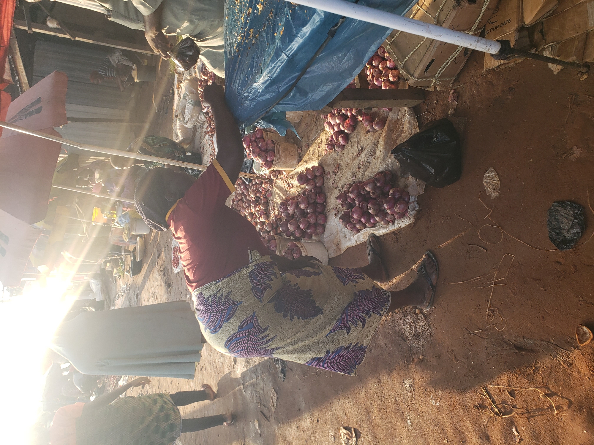 A woman, dressed in a red shirt and a patterned wrap skirt, is leaning over a table covered with a blue plastic sheet in a marketplace. She is bent at the waist, with her hands on the table. Around her, on the ground, are numerous piles of red onions, some contained in buckets. Other people and market stalls are visible in the background, with bright sunlight casting lens flare across the scene. The ground is composed of reddish-brown dirt, indicative of an outdoor market. The general atmosphere suggests a bustling commercial environment. The location is identified as Kakuri Gwari, Nigeria.