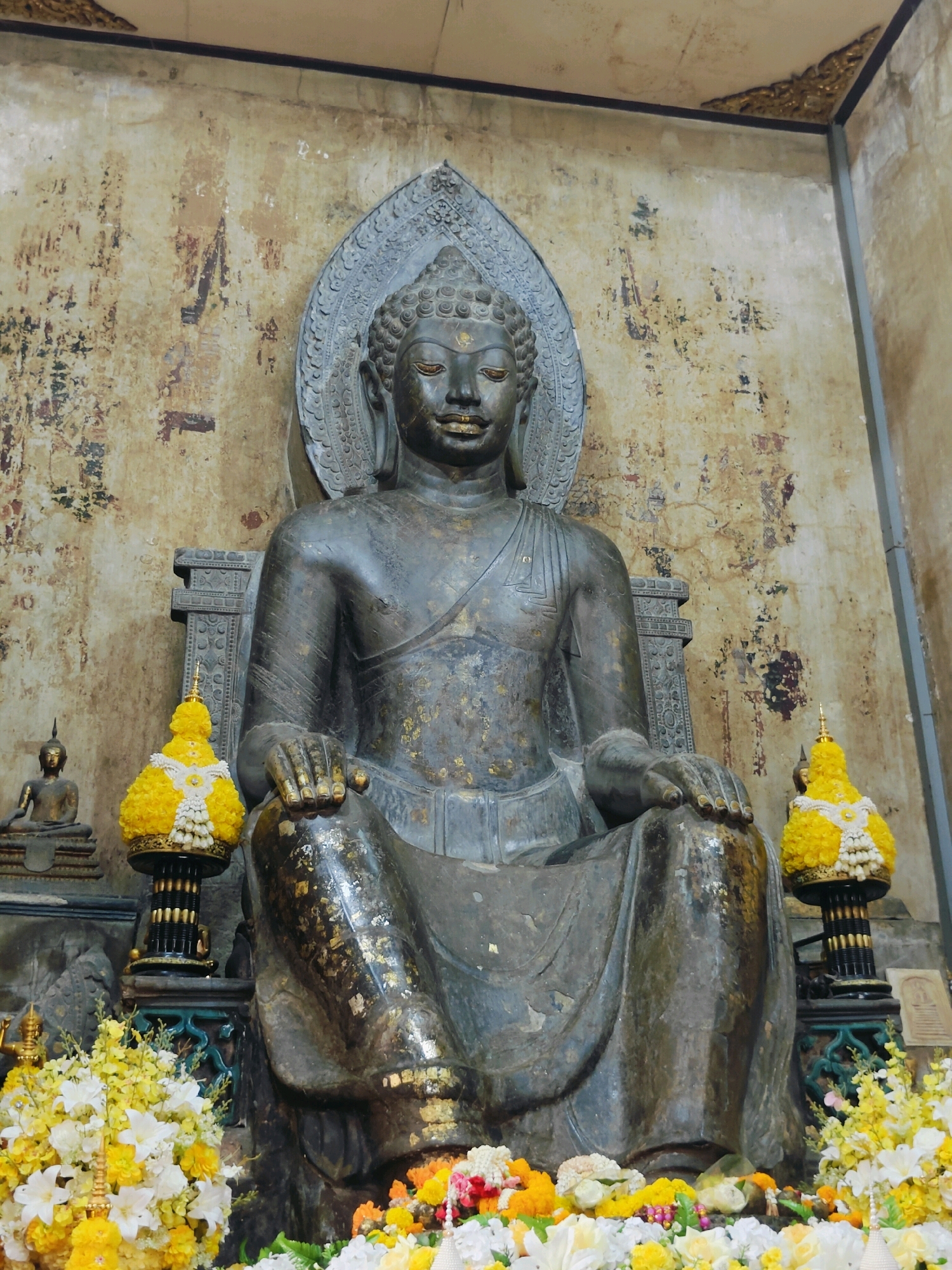 An outdoor shot depicts a large, dark bronze Buddha statue seated in meditation. The statue is adorned with gold leaf, particularly on its robe. The Buddha's hair is coiled and the urna is visible on its forehead. It wears a serene expression, with closed eyes. Behind the statue, a textured wall displays faded paintings and patches of wear. Two decorative yellow floral arrangements are placed on either side of the statue, featuring white and yellow flowers. In the background, to the left, a smaller, seated Buddha statue is visible. The foreground is filled with more vibrant yellow and white flowers, arranged in a lush display. The scene appears to be located outdoors, possibly within a temple complex or shrine in Phra Nakhon Si Ayutthaya City Municipality, Thailand, suggested by the architectural style and devotional elements.