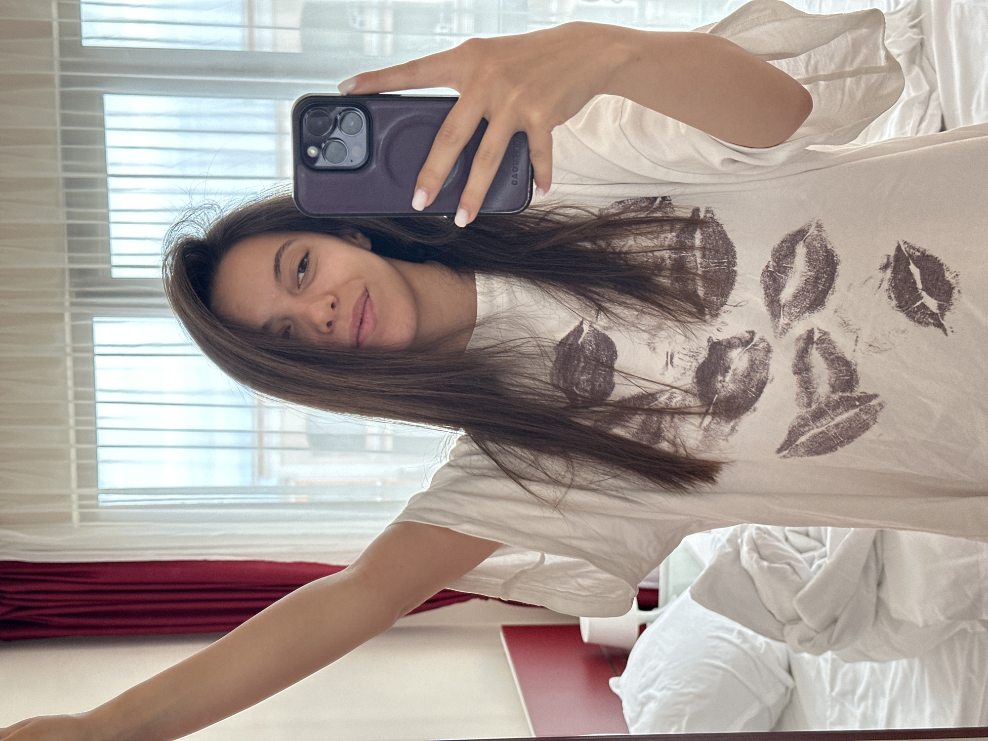 A young woman with long, dark brown hair is taking a selfie in a bedroom. She is wearing a white t-shirt with a print of dark red lipstick kisses. She is standing with her right arm raised, holding a dark purple smartphone. The background shows a bed with rumpled white linens, a bedside lamp on a red headboard, and sheer white curtains covering a window. Dark red curtains are visible on the left side of the frame. The image appears to be taken in a hotel room or residential setting in Sveti Vlas, Bulgaria.