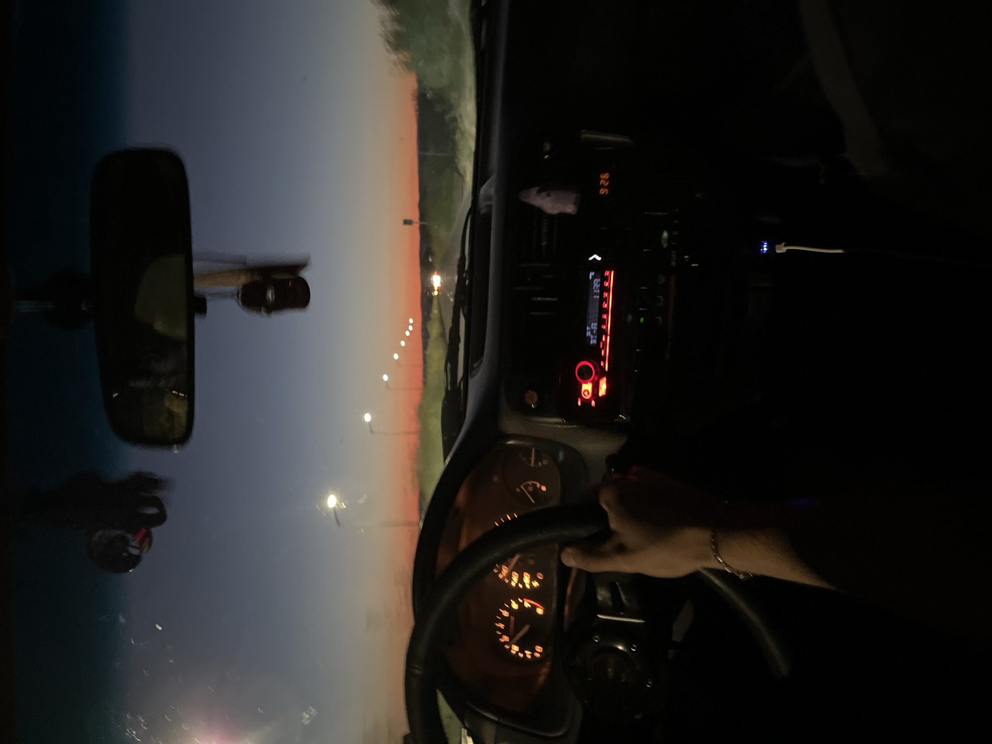 The image shows an interior view from the driver's seat of a car. A hand is on the steering wheel. The dashboard and radio are visible. The radio display shows "92.6". A USB cable is plugged in. Through the windshield, streetlights line a road. The sky is a gradient of orange and dark blue. The rearview mirror is visible. The location is not explicitly identified as Tankovo, Bulgaria.