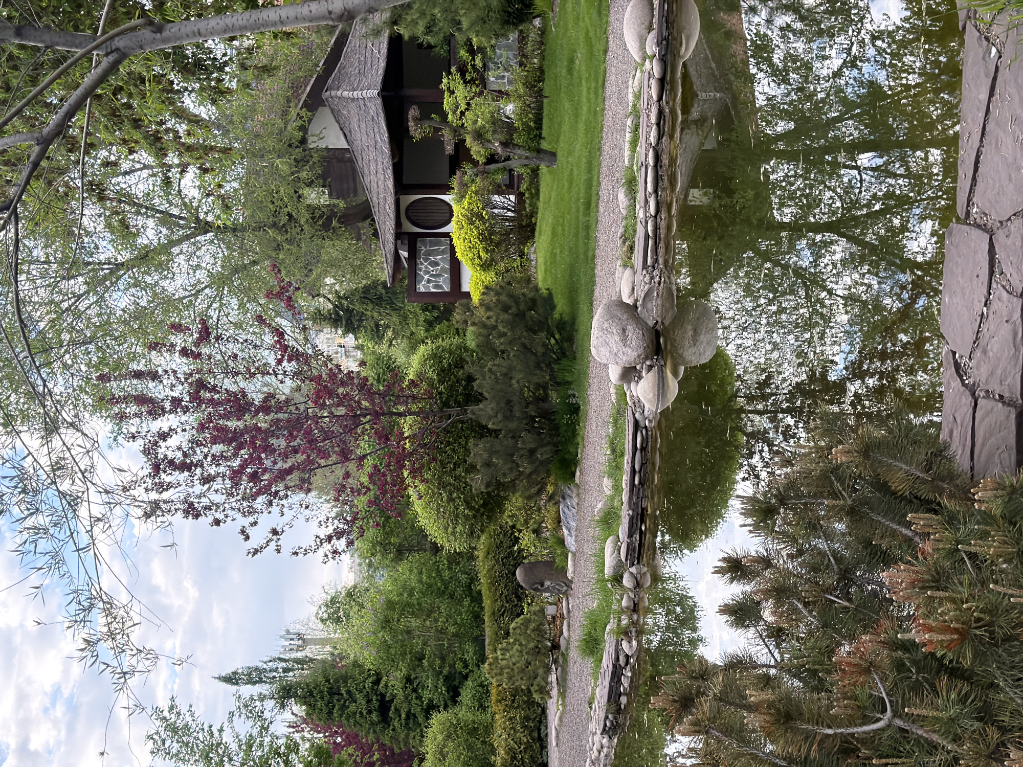 This image captures a tranquil scene in Krasnoyarsk, Russia, specifically within the city's botanical garden. The composition features a pond or lake as its central element, reflecting the surrounding foliage and sky.
Along the pond's edge, various stones and rocks create a natural border. Beyond the pond is a garden with assorted trees, shrubs, and flowers. One tree is particularly notable for its reddish-purple leaves.
A small building is visible in the distance. It has a dark roof, light-colored walls, and a round window. The building is surrounded by well-maintained greenery.
The overall atmosphere of the image is serene, suggesting a peaceful environment within the botanical garden. The water's reflection and the carefully arranged landscape contribute to the scene's aesthetic appeal.