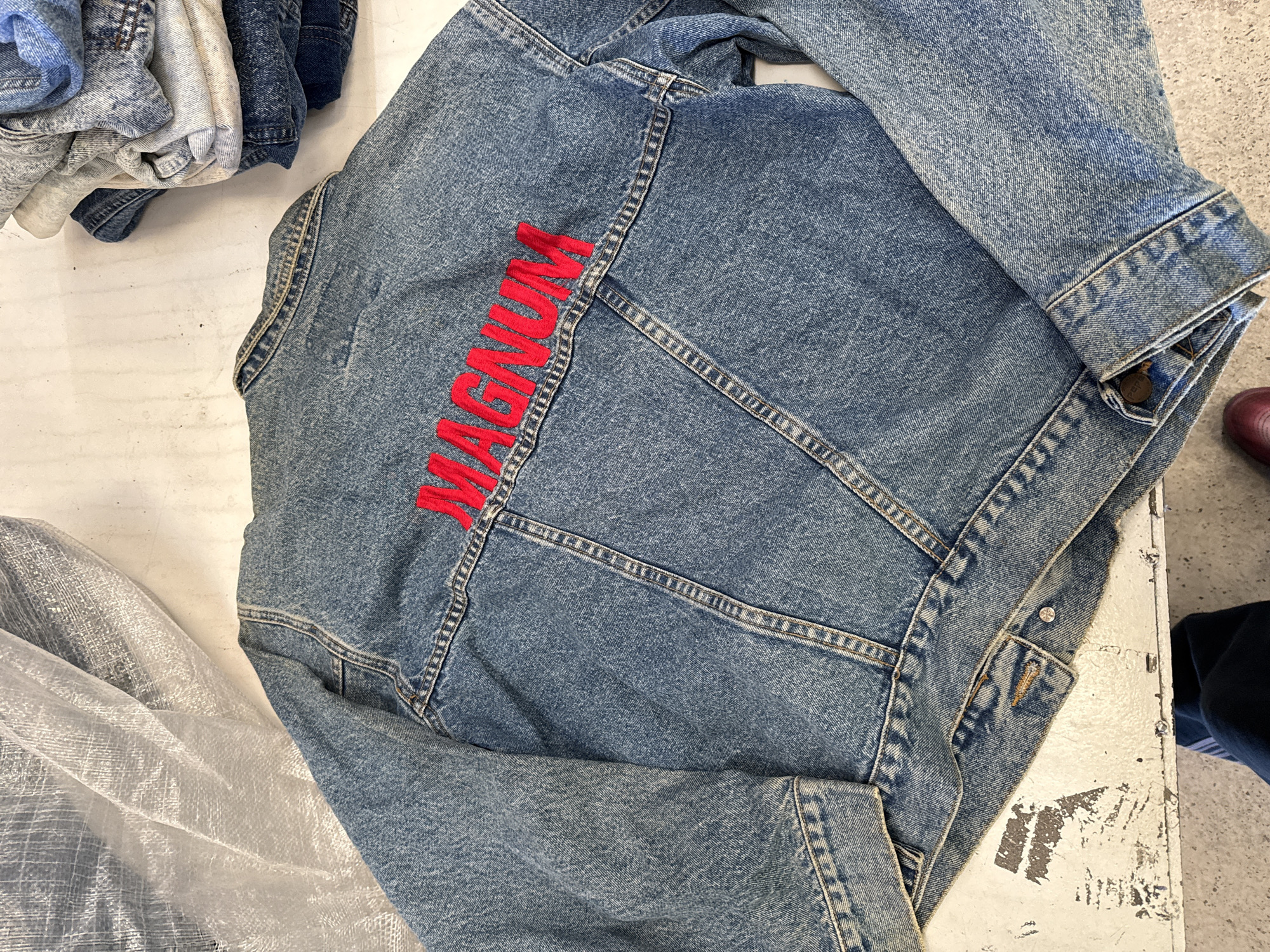 The image depicts a blue denim jacket lying on a white, possibly painted, surface. The word "MAGNUM" is embroidered in red on the back of the jacket. There are other clothes piled on the surface beside the jacket. A person is visible on the right. The location is not identifiable from the image.