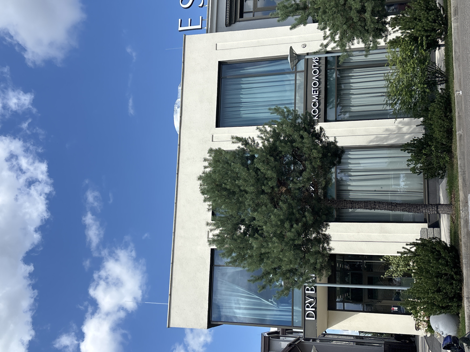 The image captures a bright, sunny day in Millennium Park, Russia, with a clear blue sky adorned with scattered white clouds. In the foreground, a light-colored, modern building stands prominently. The building features large, floor-to-ceiling windows with light blue curtains drawn inside. Above the entrance of the building, illuminated white signs are visible. One sign reads "DRY B", and another, further to the right, displays "КОСМЕТОЛОГИЯ" in Cyrillic script. Adjacent to the "КОСМЕТОЛОГИЯ" sign, a large illuminated letter "E" is visible on a separate structure, possibly part of a larger building or signage complex.
A mature pine tree with dense green foliage stands directly in front of the building, partially obscuring one of the large windows. Smaller evergreen bushes and manicured shrubs are planted along the base of the building and sidewalk. A street lamp with a modern design is positioned near the right side of the building. To the left of the main building, a portion of another building with dark siding and peaked roofs can be seen. In the lower left corner of the image, part of a grey, abstract sculpture resembling a face is visible next to some flowers and bushes. A dark-colored object, possibly a vehicle or equipment, is partially visible in the extreme lower left corner. The ground appears to be paved with asphalt and edged with a concrete curb. A person can be seen walking into the building entrance on the left side.
