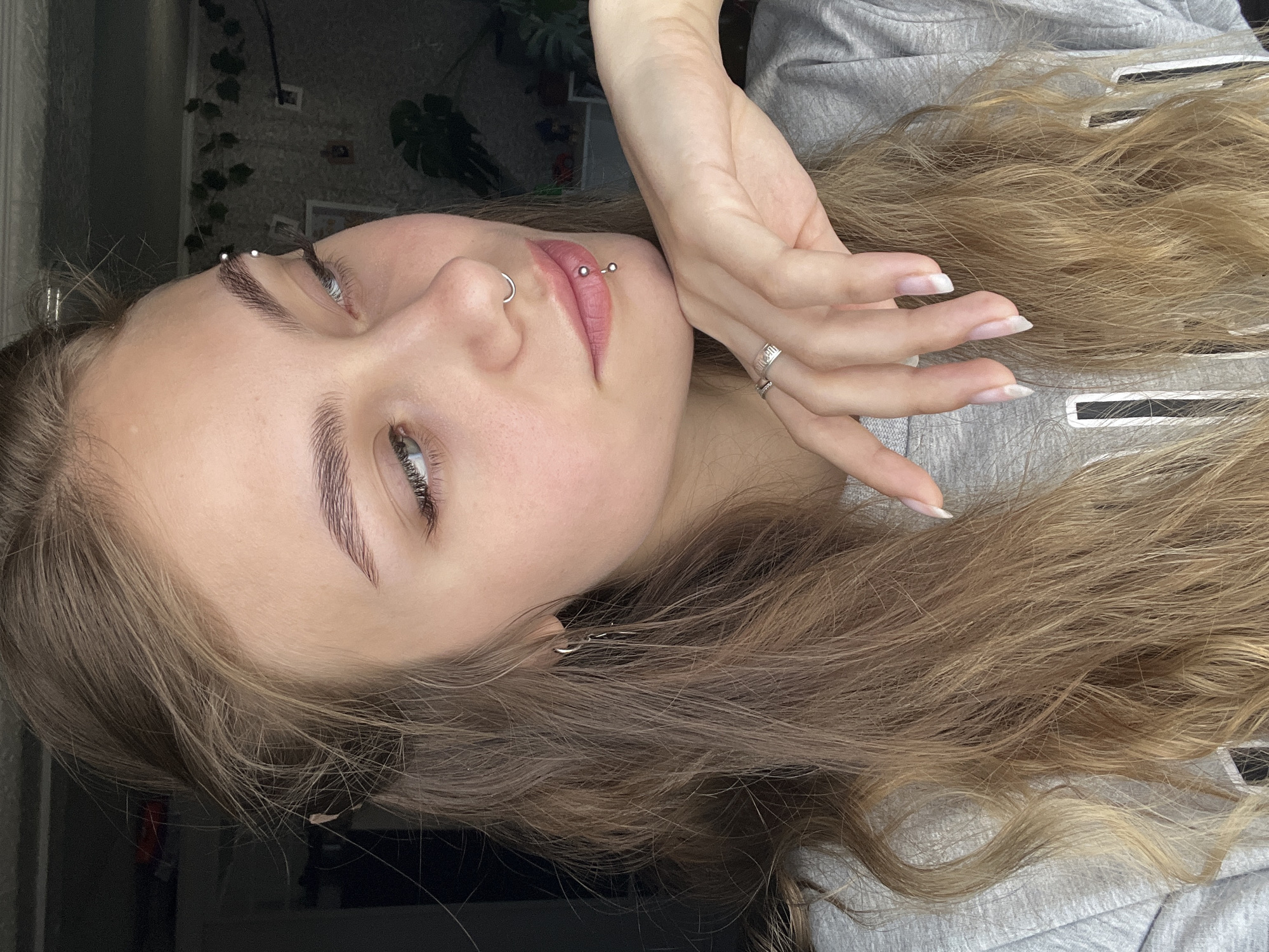 A woman with light brown hair is posing for a portrait. She is wearing a grey hooded sweatshirt. She has multiple piercings: an eyebrow piercing, a nose ring, and a lip piercing. Her nails are long and manicured with a natural-looking finish. She is wearing rings on her fingers. There is a plant in the background. The location is unknown.