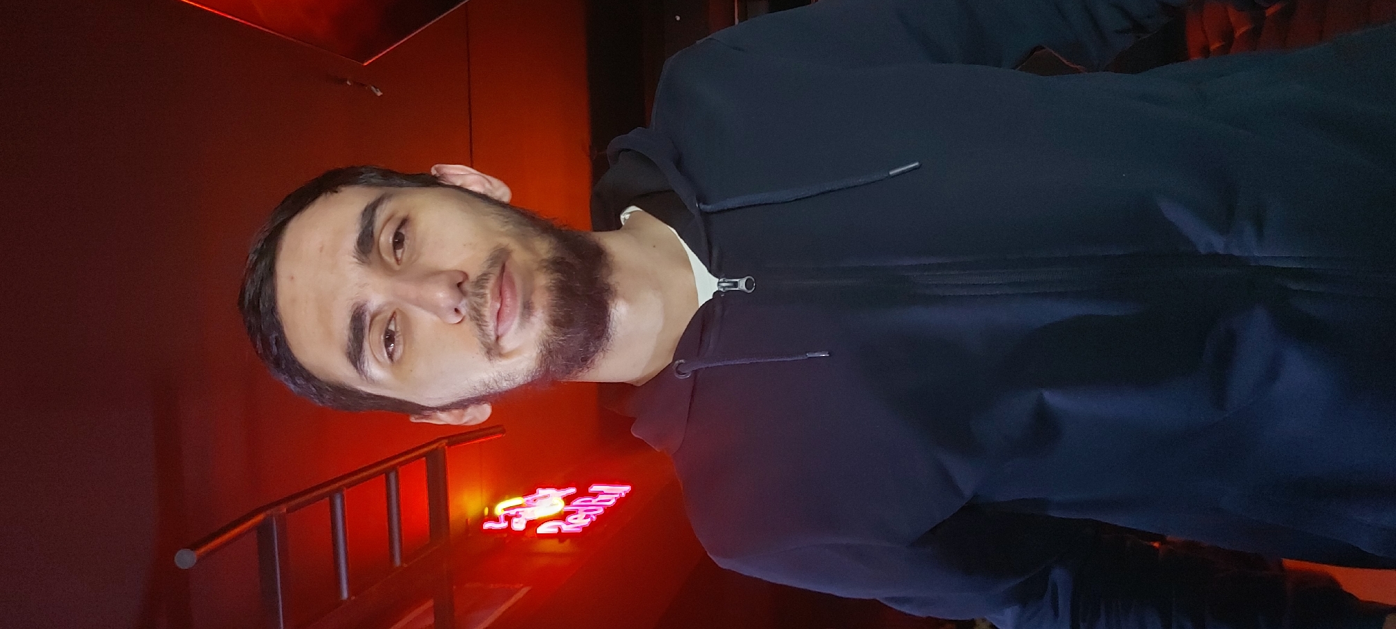 A man with short dark hair, light skin, and a beard, is facing the camera. He is wearing a dark blue hooded jacket and standing indoors. Behind him is a red-lit wall with a red neon sign that appears to read "Red Bull" in Cyrillic script. A metal handrail is visible to the left of the sign. Based on the sign and the presence of Cyrillic script, the location might be in a region like Tashkent, Uzbekistan.