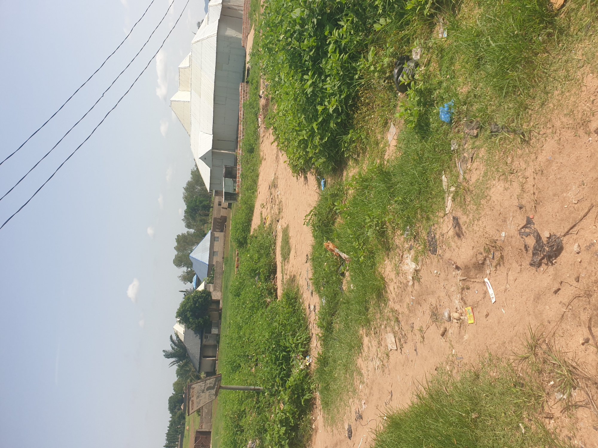 An outdoor, eye-level medium shot captures a dusty path winding through an area with overgrown green grass and scattered litter. In the background, several structures are visible. To the left, a weathered sign on a metal pole reads "WORSHIP WITH US". Beyond the sign, a chain-link fence with a gate encloses a property with a building featuring a dark, sloped roof. Adjacent to this, a smaller building with a blue, pitched roof stands. Further to the right, a large, white, multi-peaked building dominates the background, with a brick border in front of it. Trees and other vegetation are scattered throughout the scene, and overhead, three power lines stretch across the pale blue sky, which is dotted with a few wispy clouds.