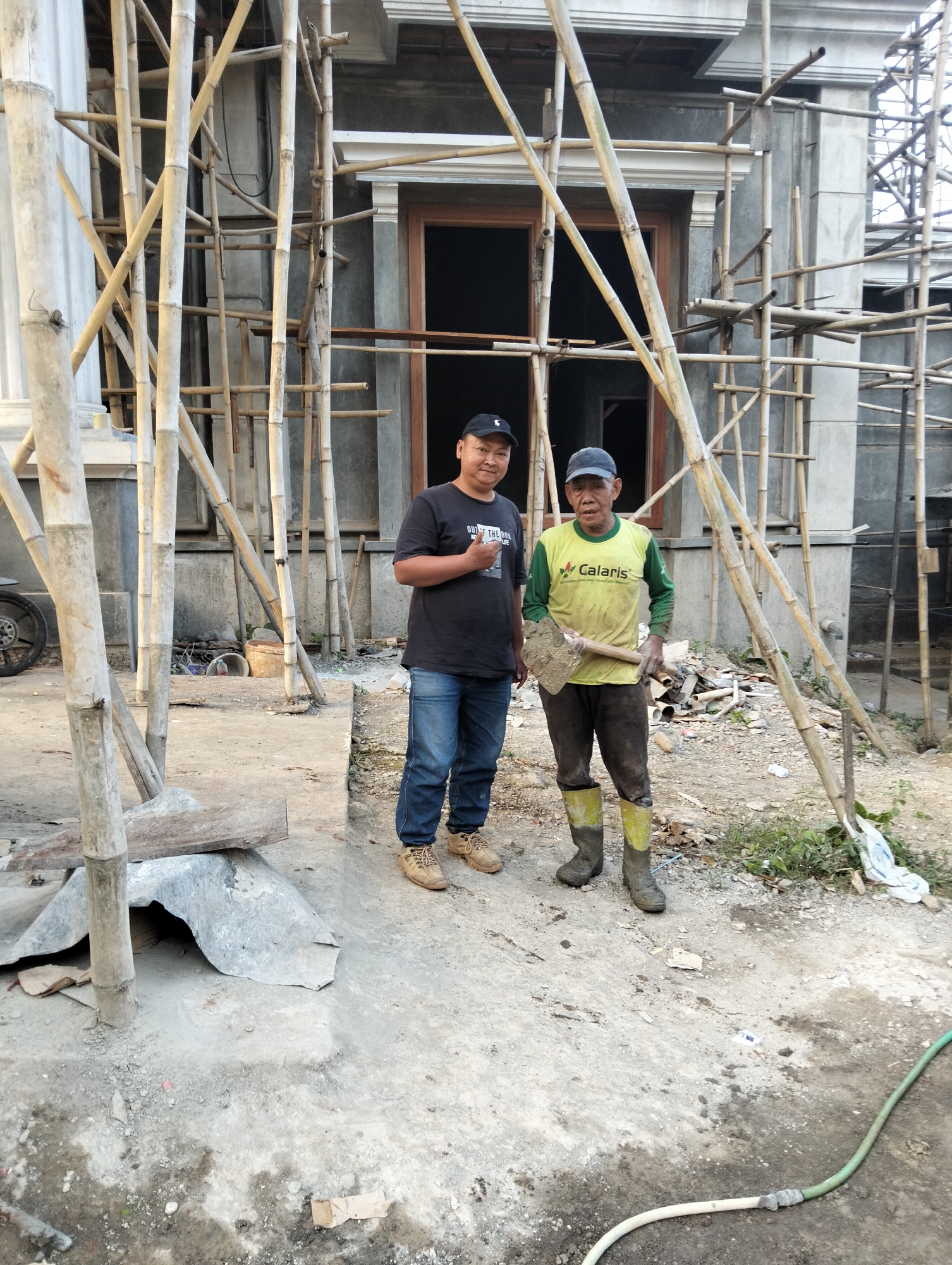 Two men are standing outside a building under construction in Durenombo, Indonesia. The man on the left is wearing a black t-shirt, blue jeans, and brown work boots. The man on the right is wearing a green and yellow long-sleeved shirt, dark pants, and yellow rubber boots. He is holding a shovel. Both men are facing the camera. The building behind them is unfinished and is supported by bamboo scaffolding. The ground is dirt and gravel.