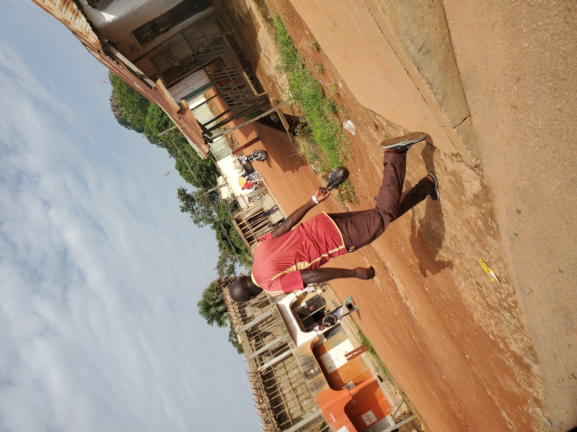 The image depicts a man in a red shirt and brown pants walking away from the camera in a dusty, outdoor urban environment. The man is carrying a black bag in his right hand. In the background, there are several buildings, some appearing to be under construction with exposed wooden framing, and others with corrugated metal roofs. A woman is visible walking in front of one of the buildings. A motorcycle is parked nearby. The ground is covered in reddish-brown dirt and some scattered debris. The sky is cloudy. The scene is consistent with a street in Bukuru, Nigeria.