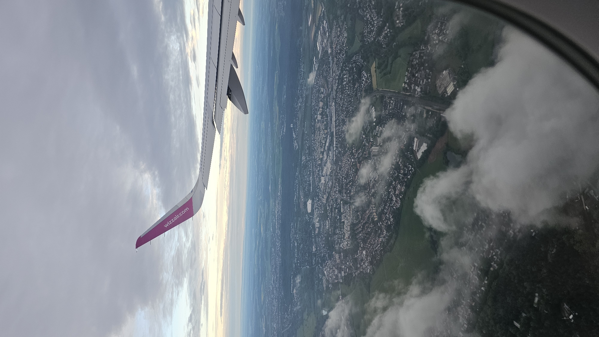 An oblique aerial view captured from an airplane window shows a Wizzair aircraft wing over the cityscape of Fürth, Germany. The wing, colored in white with a pink tip bearing the "wizzair.com" logo, extends to the upper left of the frame against a cloudy sky. Below, the urban landscape is visible, consisting of densely packed buildings interspersed with green spaces, parks, and patches of vegetation. The presence of a highway can be observed cutting through the city, indicating transportation infrastructure. Clouds obscure some portions of the cityscape. The image also captures the curvature of the airplane window, adding a subtle distortion to the overall view.
