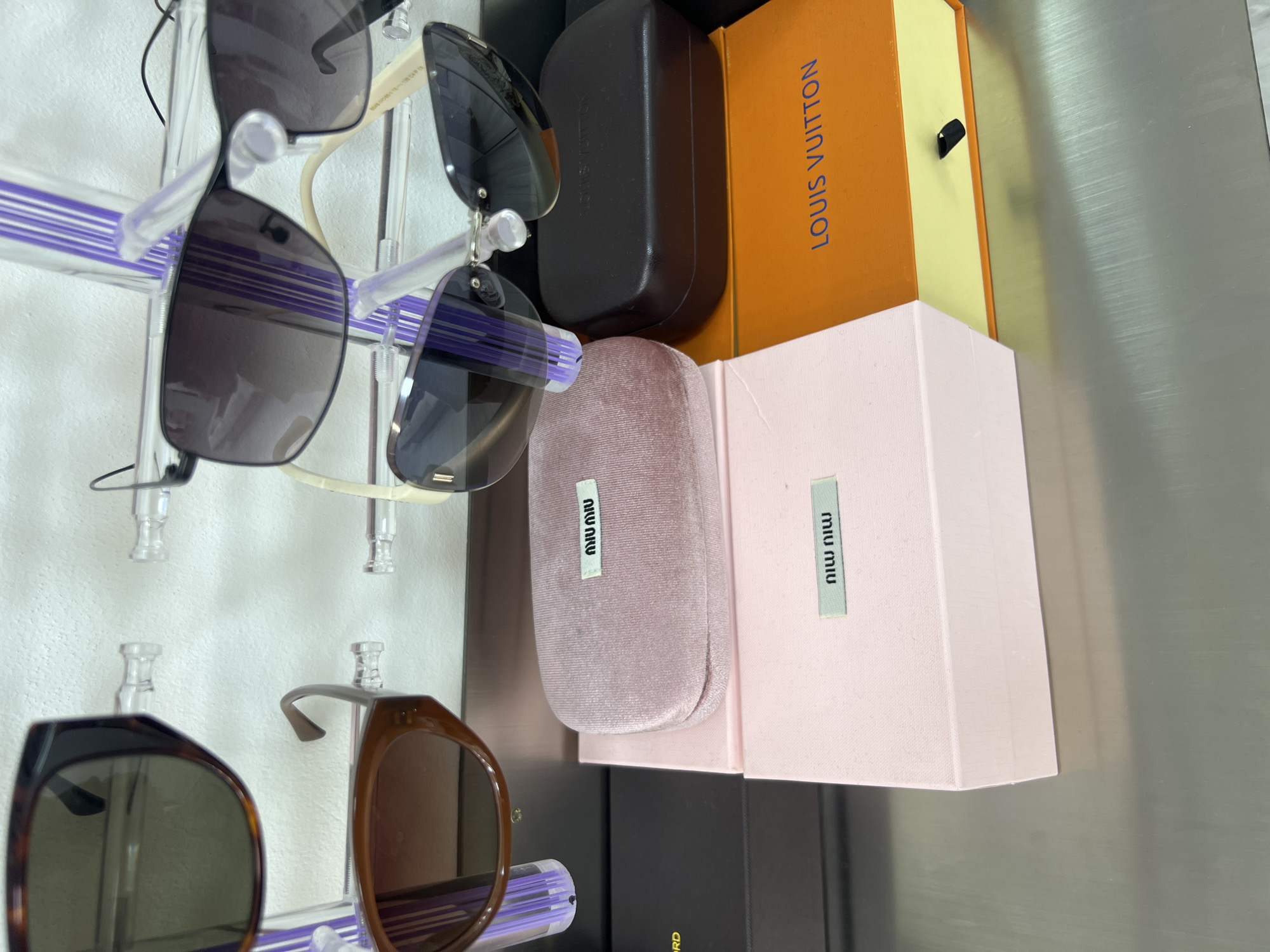 The image shows a close-up of several pairs of sunglasses on display. The sunglasses are of various styles and colors, and some are displayed in their cases. In the foreground, there are two pink boxes, one with a velvet texture and the other plain. Both boxes have a small label with the "MIU MIU" logo. To the right of the pink boxes is an orange Louis Vuitton box with the brand name printed in white. Behind the boxes, more sunglasses are visible, some mounted on clear plastic stands against a white wall. The overall setting appears to be a retail display, possibly in a store. The location of Osh City, Kyrgyzstan is not discernible from the image.