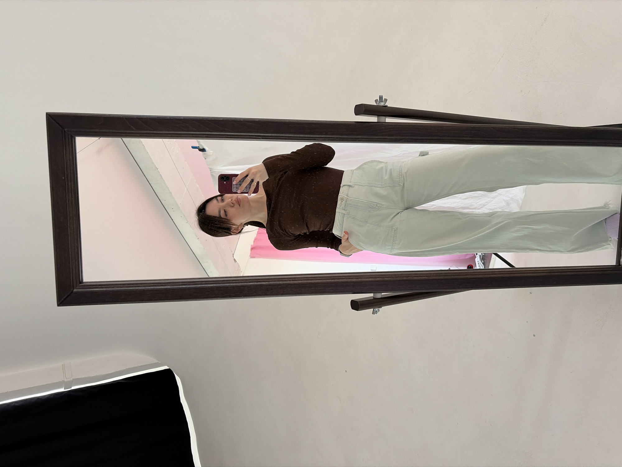 A young woman takes a mirror selfie. She wears a sheer brown long-sleeved top and light blue jeans. The mirror has a dark brown frame and rests on a stand. The background reflects a pink wall. The location may be Chaikovsky, Russia.