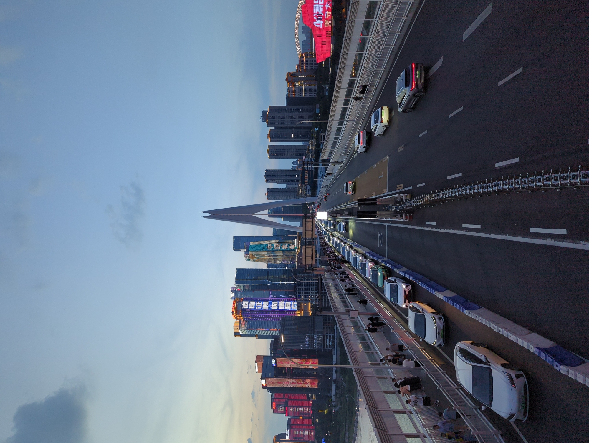 The image shows a cityscape at dusk, featuring a wide bridge with a multi-lane roadway and a pedestrian walkway. On the bridge, there is a traffic jam with numerous cars moving in the same direction. Several pedestrians are visible walking on the walkway. In the background, a modern city skyline is illuminated by colorful lights emanating from tall buildings. One prominent structure on the bridge is a large, arched support. The sky is a mix of blue and gray clouds, indicating the transition from day to night. The location is identified as Yuzhong District, China.