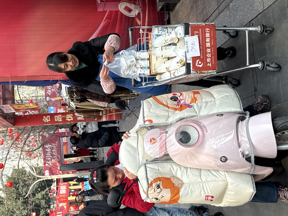 An outdoor scene shows a woman in a black jacket putting items from a shopping cart into a plastic bag. The cart is filled with bottles and has a sign advertising 30 yuan from the Bank of Chongqing. A girl, bundled in a cartoon-themed blanket and riding on a scooter, is visible in the foreground. The background is a bustling street with red banners and decorations. The text on the banners includes "Xin Chun Ji" and "Mingpin You".
