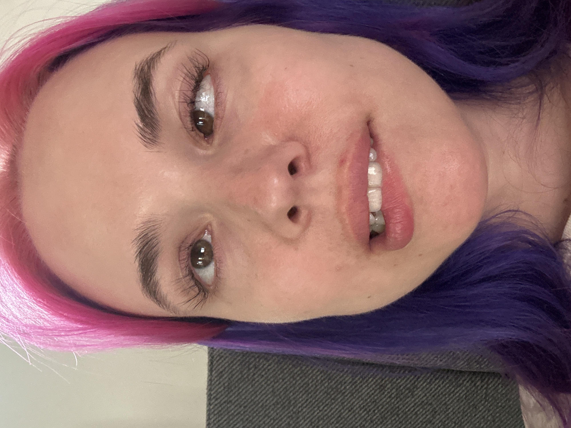A close-up, tilted selfie of a woman with pink and purple hair. She has brown eyes and is smiling slightly, showing her teeth. Her skin tone is light, and she appears to be indoors. There is a gray patterned object in the lower part of the frame. There is no indication of this image being located in Zagreb, Croatia.