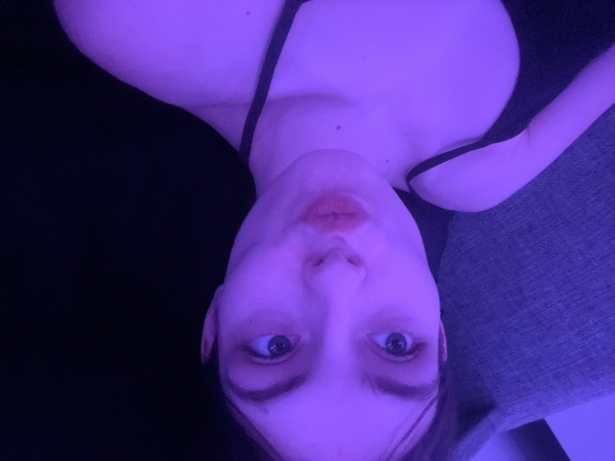 A close-up, inverted perspective photograph depicts a person with fair skin illuminated by a strong purple light. The person is wearing a black top with thin straps. The background consists of dark and gray areas, potentially indicating furniture or a wall. The image gives no indication of location, particularly Saint Petersburg, Russia.