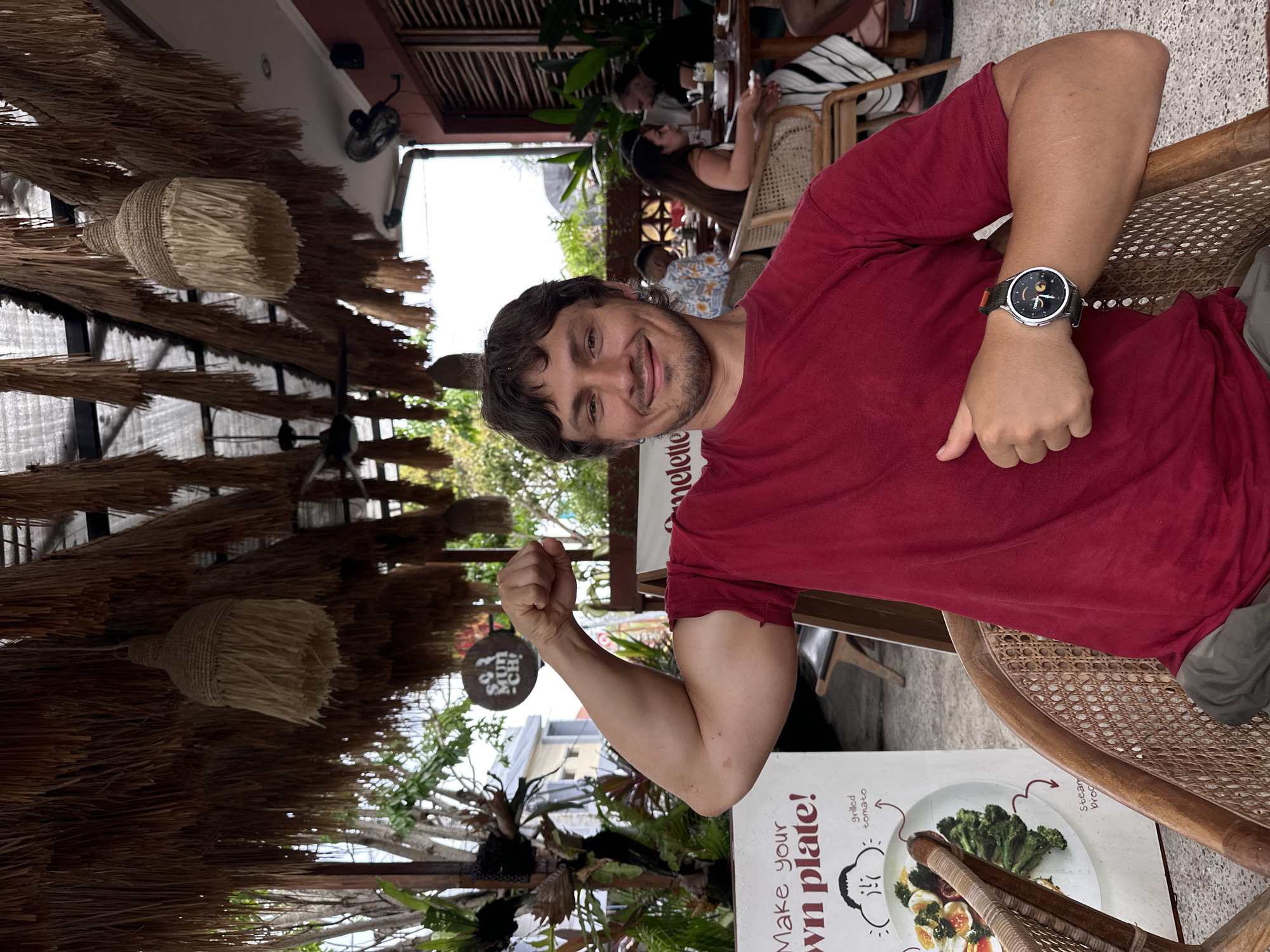 A young adult male, with brown hair and stubble, wearing a red t-shirt and grey pants, is seated in a wicker chair at an outdoor cafe. He is flexing his bicep, with his left arm raised. He is smiling and giving a thumbs-up with his right hand. In the background, other patrons are seated at tables. The cafe is covered by a thatched roof, and tropical plants are visible. A sign behind the man reads "Make your own plate!". This scene is located in North Kuta, Indonesia.