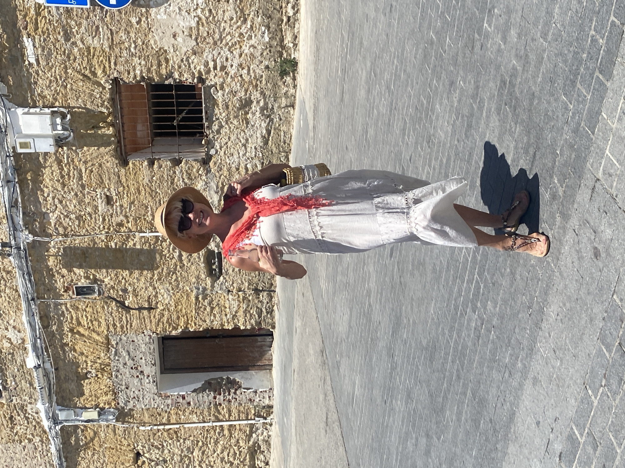 A woman stands on a cobblestone street in front of an old stone building in Conil De La Frontera, Spain. The building has a rough, textured facade with a wooden door on the left and a barred window on the right. Electrical conduits and boxes are visible on the exterior of the building. The woman is wearing a white, tiered dress, a straw hat, sunglasses, and sandals. She is holding a bottle of water in her right hand and has a woven bag on her right shoulder. A red scarf is draped around her neck. The lighting suggests it is a sunny day.