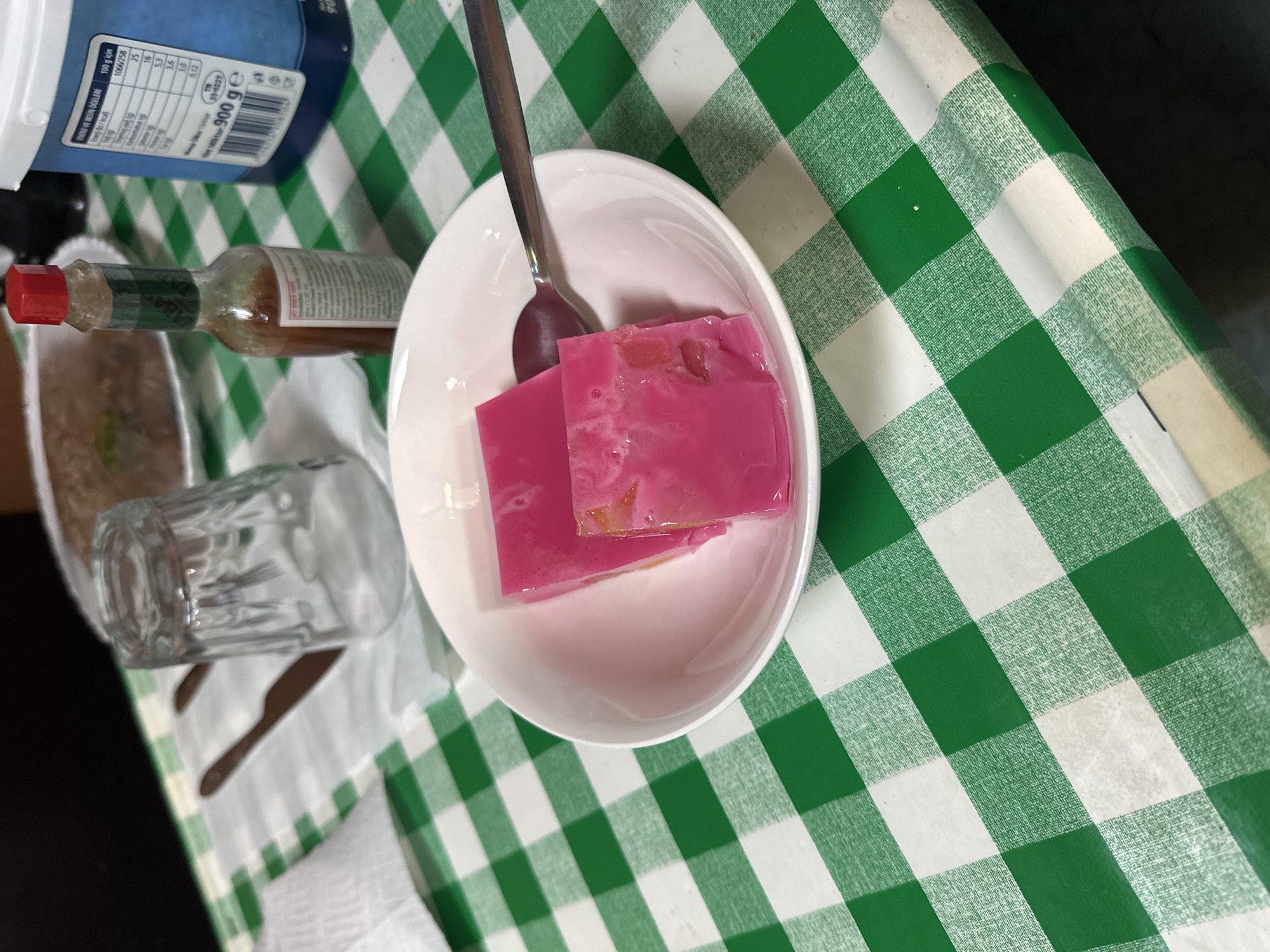 Here is a forensic description of the image:
The image shows a tablescape on a green and white checkered surface. A white bowl contains two rectangular blocks of pink gelatin dessert, served with a silver spoon. To the left of the bowl is a small glass tumbler, a partially visible plate with food remnants, a bottle of hot sauce with a red cap, and a larger white container with a blue label and visible nutrition facts. The table appears to be situated indoors, with the checkered surface suggesting a table cloth or placemat.