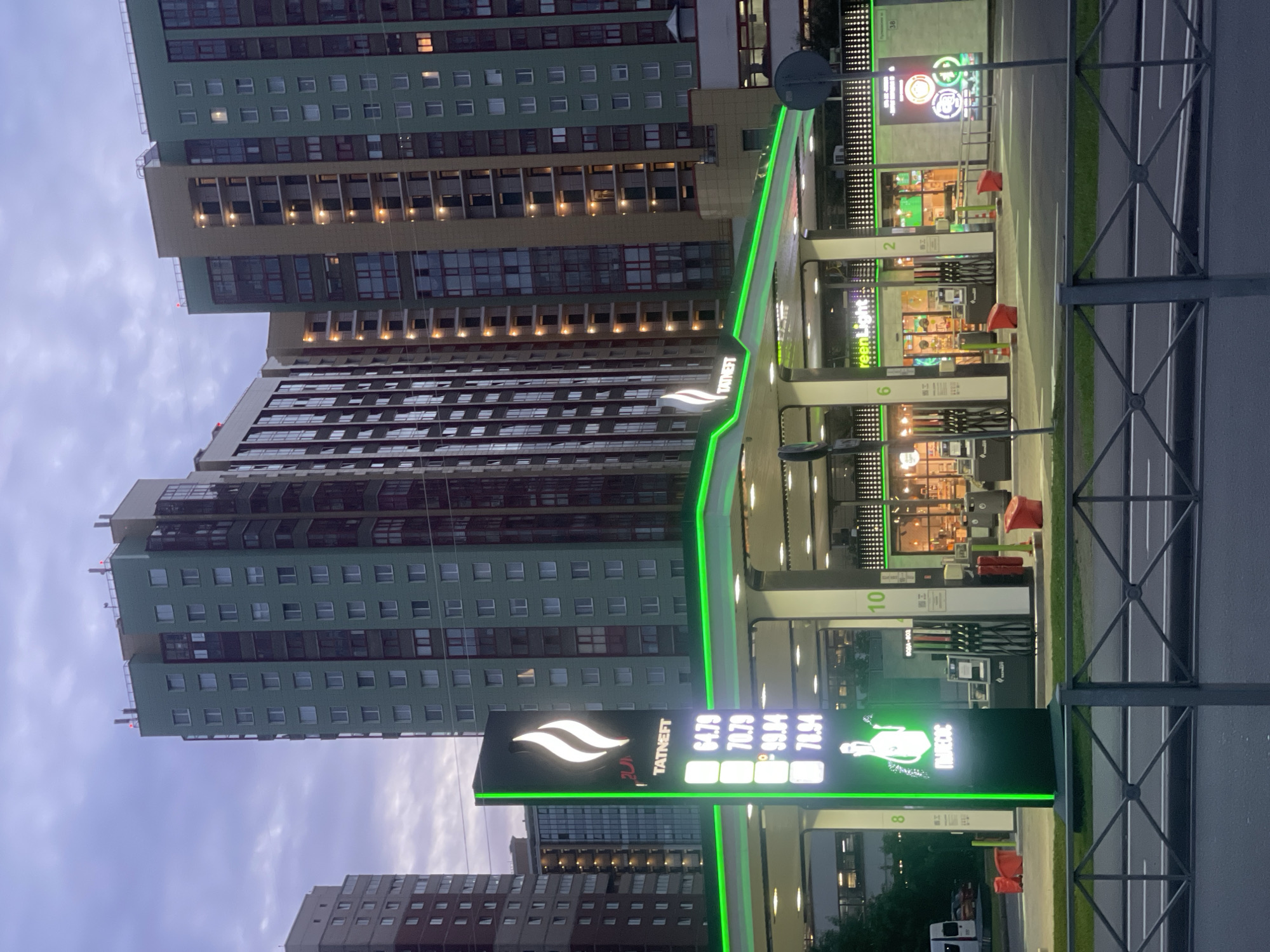 The image shows a modern gas station at dusk, with several tall residential buildings in the background. The gas station features a prominent sign with the "TATNEFT" logo and fuel prices displayed. Several fuel pumps are visible, and the canopy of the gas station is illuminated with green LED lights. In the foreground, a metal fence separates the road from the gas station area. The sky is overcast, indicating the time of day is either early morning or late evening. There are no people visible in the scene.