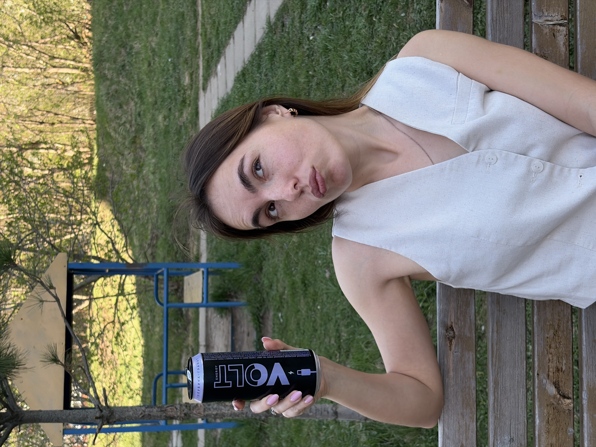 A woman is holding a black and purple Volt energy drink can while sitting on a wooden bench. She is wearing a white, button-down sleeveless top and a delicate necklace. The scene appears to be outdoors, with grass and greenery visible in the background, and what looks like playground equipment nearby. The energy drink can has the word "VOLT" written in large, purple letters and a small icon resembling a charging plug. The can also displays text in Russian, including "ГОЛУБИКА-ГРАНАТ."