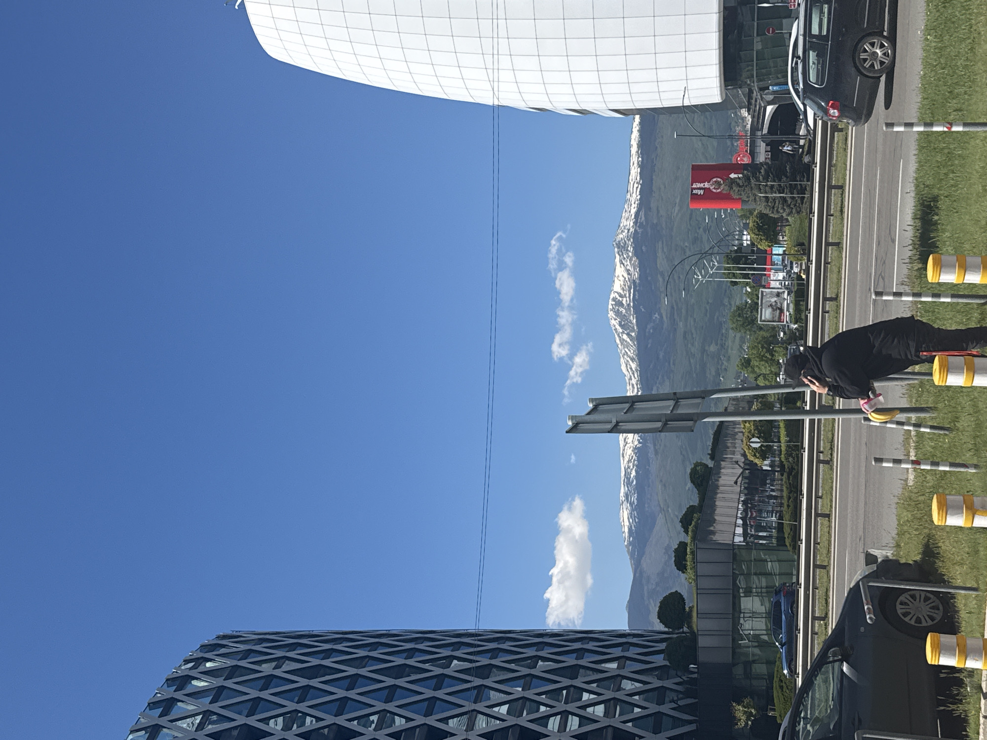 The image depicts an outdoor scene in Sofia, Bulgaria. A clear blue sky dominates the upper portion of the frame, juxtaposed with snow-capped mountains in the background. In the foreground, two modern buildings are visible: one with a white, grid-like facade, and the other with a darker, diamond-patterned glass exterior. A multi-lane road runs through the center of the image, lined with greenery, streetlights, and striped yellow and white bollards. Cars are parked and moving along the road. A person wearing black clothing is seen near the bollards. A red billboard and other signage are visible in the distance.