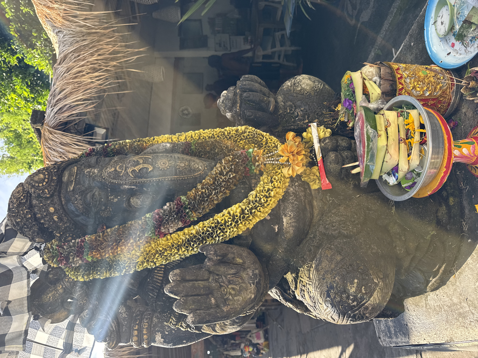 The image, rotated clockwise, captures a large, dark stone statue adorned with garlands and surrounded by traditional offerings, set in an outdoor or semi-outdoor environment in Kuta Selatan, Indonesia.
The central subject is a substantial, weathered dark stone statue, possibly representing a deity like Ganesha, given its rounded form and visible hands. The statue is elaborately decorated with a long, thick garland of bright yellow elements, possibly made from flowers or paper, draped across its body. A second, shorter garland with multicolored elements is also visible. A black and white checkered cloth, known as a poleng, is draped over a section of the statue's left side, a common symbol in Balinese spiritual contexts.
Positioned near the statue are several traditional Balinese offerings. These include various vessels and bowls filled with fresh leaves (likely banana or palm leaves), vibrant purple and yellow flowers, and other ritualistic items, commonly known as canang sari. One cylindrical offering container is particularly ornate, decorated with intricate red and gold patterns. A blue and white enamel plate, also holding offerings, is present.
The background shows elements of Balinese architecture, including a thatched roof structure and lush green foliage from trees. The scene is bathed in bright sunlight, which creates strong shadows and distinct lens flares across the statue, indicating a midday or early afternoon setting. The overall composition strongly suggests a sacred site or shrine, reflecting ongoing religious and cultural practices specific to Bali. No visible text is present in the image.