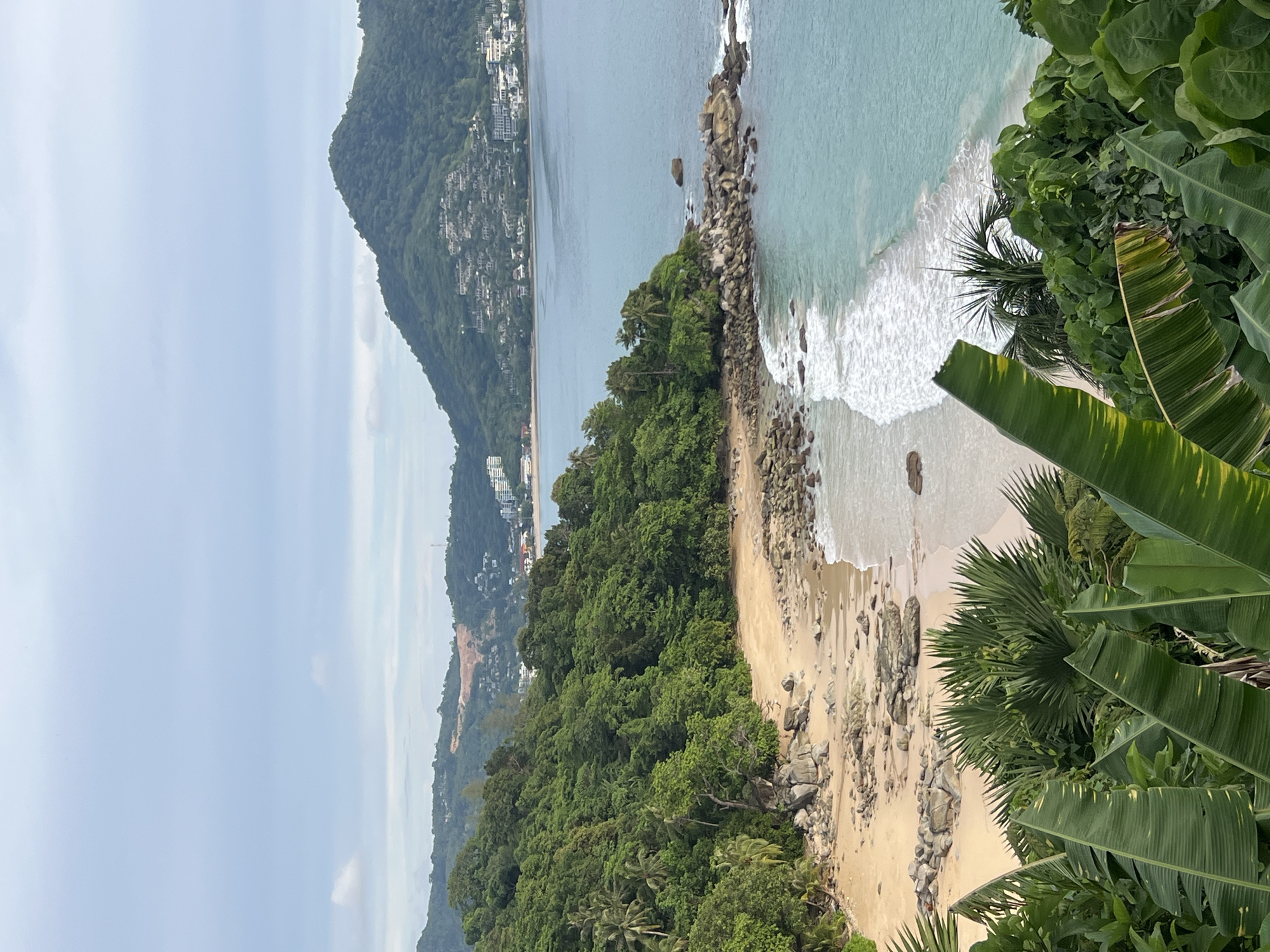 The image captures a scenic view of a secluded beach in Ban Bang Thao, Thailand. Lush green tropical foliage, including large banana leaves and palm trees, frames the foreground. The beach itself is sandy with scattered rocks and boulders, leading to clear turquoise waters with gentle waves. In the background, a verdant, tree-covered hill rises, dotted with buildings that suggest a coastal settlement. The sky is overcast with a soft, diffused light, indicating a cloudy day. There are no people visible in the image.