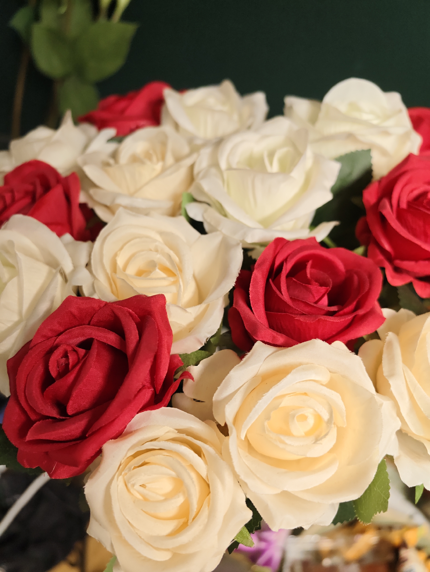 A close-up, overhead shot captures a vibrant bouquet of artificial roses, alternating between deep red and creamy white. The roses are densely packed, with their layered petals unfurling in classic spiral patterns. A few dark green leaves are visible peeking through the blossoms, providing a contrast to the floral colors. The background is a dark, solid green, creating a dramatic setting that emphasizes the flowers. The lighting is focused on the bouquet, casting soft shadows and highlighting the texture of the petals. The image appears to be a still life composition, likely an indoor setting. There is no indication of the city of Petah Tikva, Israel in the image.