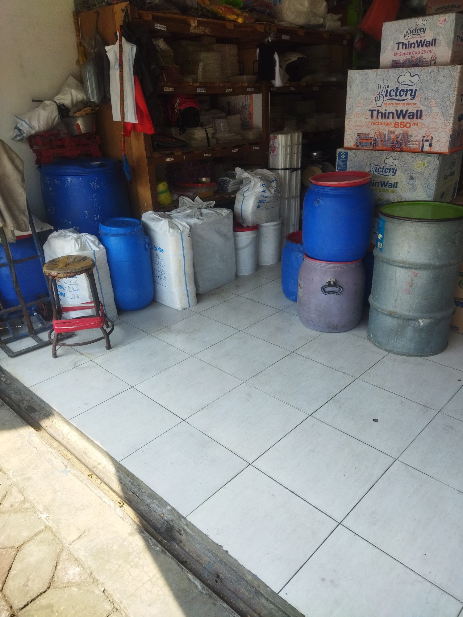 The image depicts the interior of a storage or retail space, likely a shop, in Giripurwo, Indonesia. The environment is cluttered with various goods. Dominating the foreground are several large plastic and metal barrels of different colors, including blue, purple, and gray. Some of these barrels are stacked. In front of the barrels, a small wooden stool with a red band around its legs is visible. To the left, part of a metal shelving unit is present, with more barrels and bags.
Behind the barrels, shelves are stocked with boxes and bags. Prominently displayed are several boxes of "Victory ThinWall" products, suggesting they are food containers or similar items, with different volumes indicated (e.g., 25 ml, 650 ml, 2000 ml). Other boxes and bags, some white and some transparent, are also on the shelves.
The floor is tiled with square white tiles. In the background, wooden shelving units are filled with an assortment of items, including what appear to be plastic sheeting rolls, bags, and other packaged goods. A red and white striped broom is propped up against one of the shelves. The lighting suggests an indoor environment with some natural light entering from the side, casting shadows on the floor. The overall impression is one of a busy, functional storage area.
