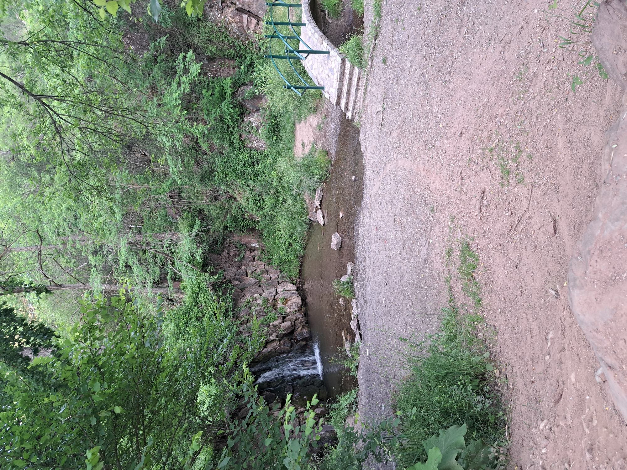This media file depicts a scenic natural setting in Lokorsko, Bulgaria. The image is oriented vertically and captures a small waterfall cascading over rocks into a stream. Lush green trees and vegetation surround the water, creating a vibrant, natural environment. A pathway, composed of gravel and dirt, runs alongside the stream, leading to a stone staircase and a small arched bridge with green railings. The time of day appears to be daytime, and the weather seems overcast or shaded, contributing to the soft lighting. The scene evokes a sense of tranquility and the beauty of nature, possibly a park or a hiking trail. There are no visible people or text in the image.