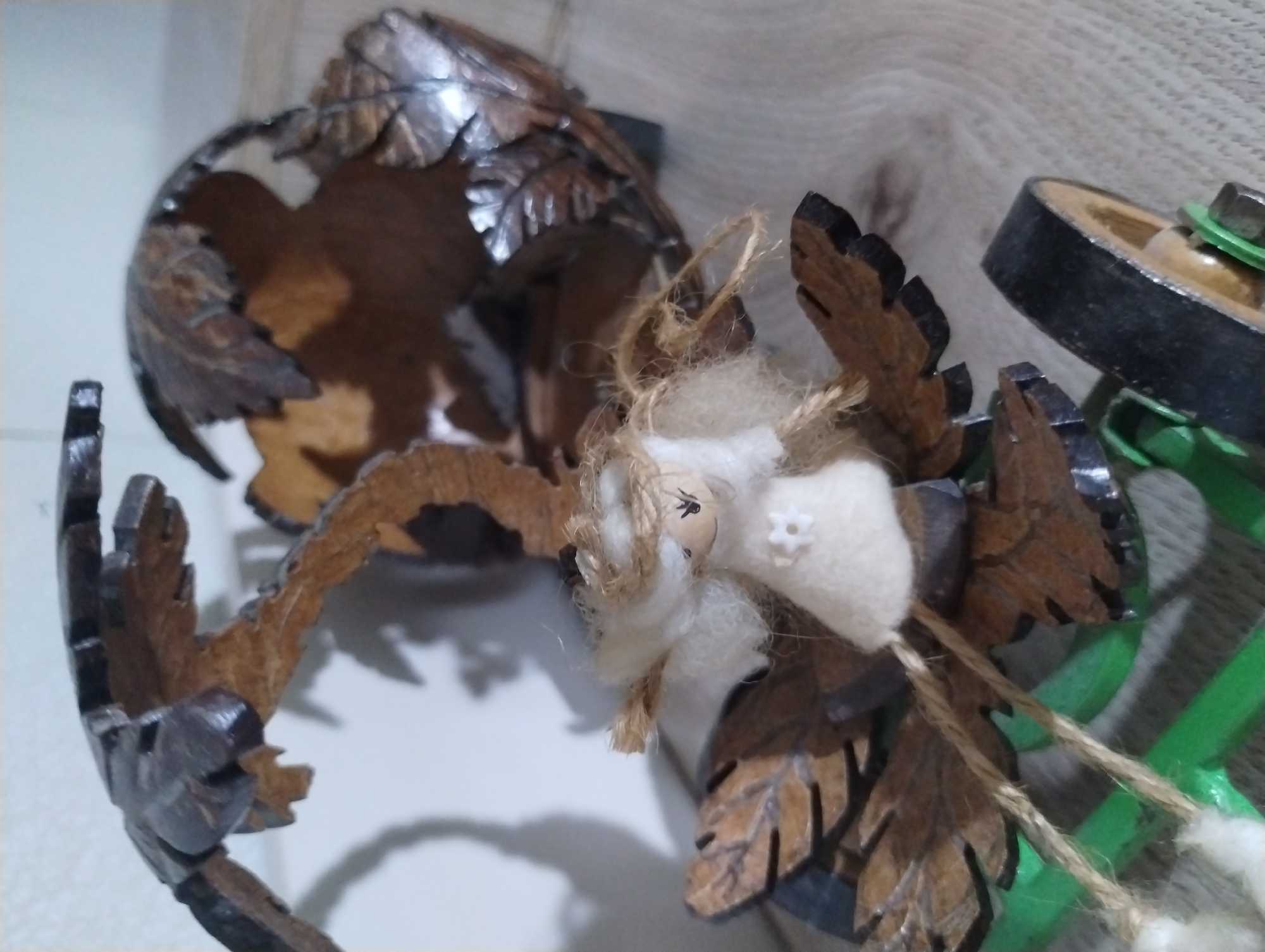 This close-up image captures a small, handcrafted doll or figurine nestled among decorative wooden elements. The doll, centrally located, features a light-colored, possibly felt, head with two simple dark dots representing eyes and a tiny white star-shaped embellishment. It has soft, fluffy, light-colored material, likely cotton or wool, forming its hair or body, from which two strands of natural-colored jute or twine hang, each terminating in a small, fluffy tuft.
The doll is surrounded by intricate, dark brown wooden carvings shaped like leaves, some appearing polished or varnished with a subtle sheen. These wooden pieces seem to be part of a larger, rustic, or nature-themed decorative structure. In the background, particularly to the right, a segment of a green object, possibly a mechanical or decorative piece, is visible, along with a metal bolt and a dark wooden wheel-like component. The lighting suggests an indoor setting. No specific activity or event is taking place, and there are no visible texts. The scene is a static arrangement of decorative items. The location context is Roza, Bulgaria.