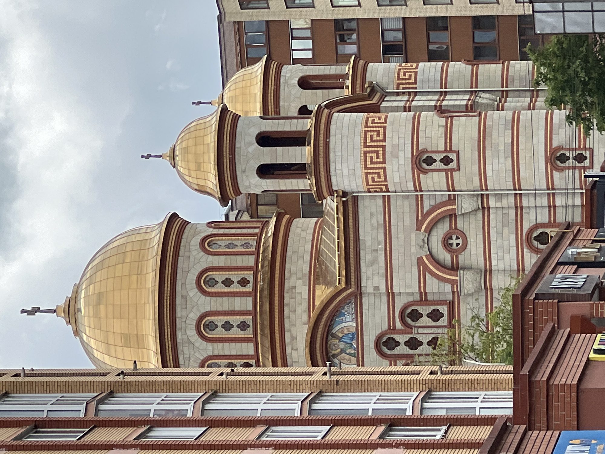 The image depicts a section of the Cathedral of the Ternopil Eparchy of the Ukrainian Orthodox Church in Ternopil, Ukraine. Visible are the golden domes, the unique brickwork pattern adorning the building, and an ornate architectural design. The church is located amongst other buildings, including an adjacent building with windows that suggest a multi-story structure. The sky is overcast.
