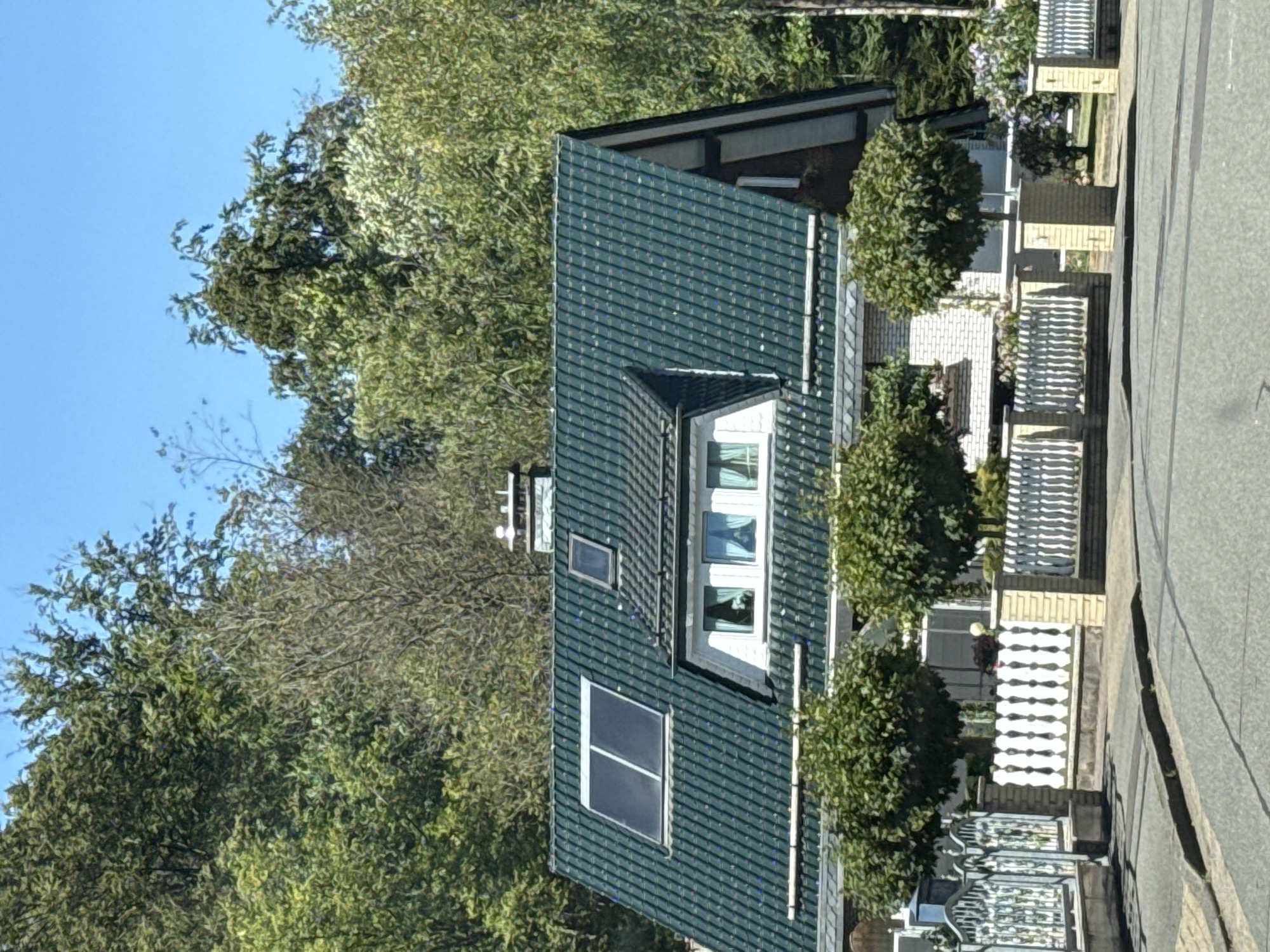 The image captures a detached house with a dark green tiled roof, situated in the city of Bad Lauterberg im Harz, Germany. The house features a dormer window and solar panels integrated into the roof. A white picket fence with brick pillars encloses the property, and several manicured trees are visible in the foreground. Behind the house, a dense forest of mature trees forms a natural backdrop under a clear blue sky. The scene appears to be a residential street, with a paved road and curb in the foreground. No people are visible in the image.