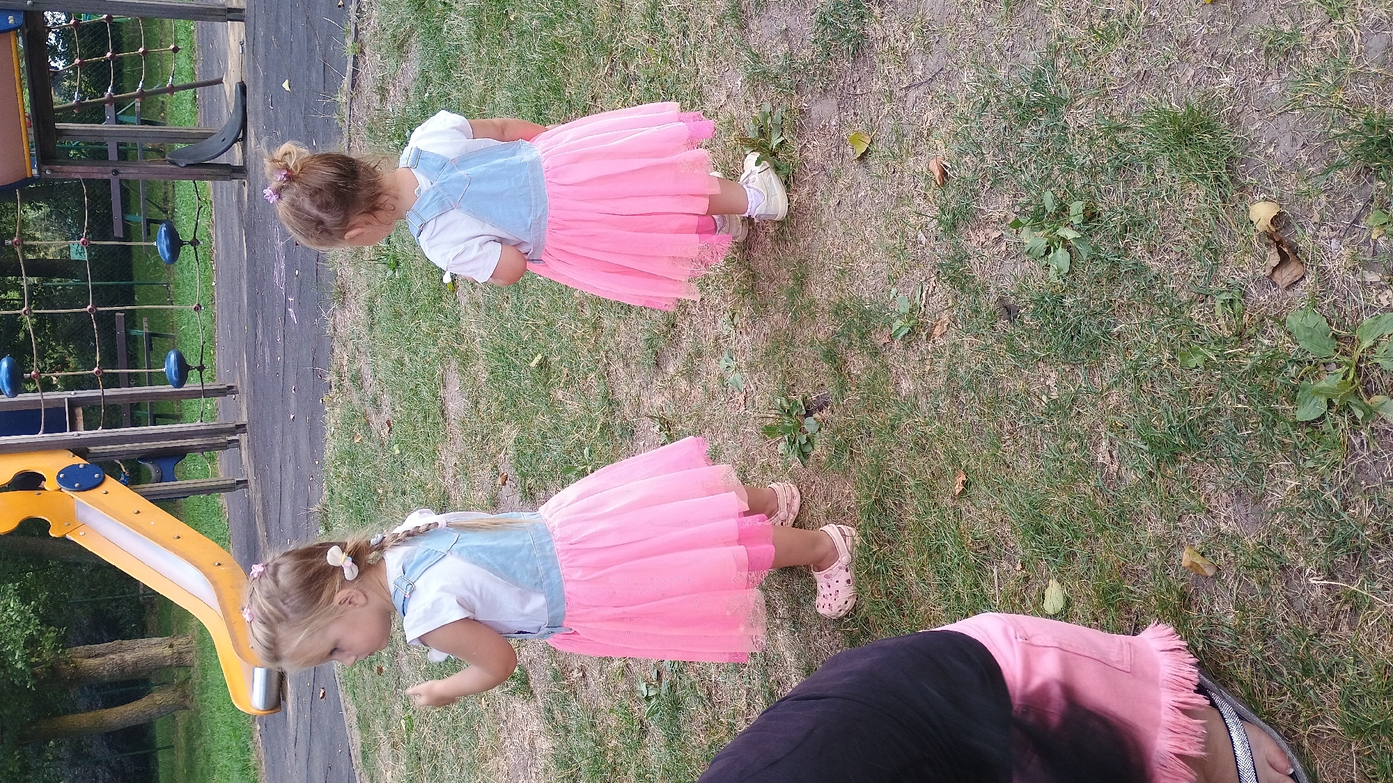 In Sokolov, Czechia, two young girls stand on a grassy area in a playground. The girl on the left, facing left, has blonde braided hair adorned with pink flowers and wears a white t-shirt with blue denim overalls and a pink tutu. She is wearing light-colored clogs. The girl on the right, facing away from the camera, also has blonde hair tied in a bun with pink bows and wears similar attire, including a white t-shirt, blue denim overalls, and a pink tutu, with white shoes. A person's legs, dressed in black and pink, are visible in the lower left corner of the frame. In the background, a colorful yellow and blue playground slide and climbing structure are present.