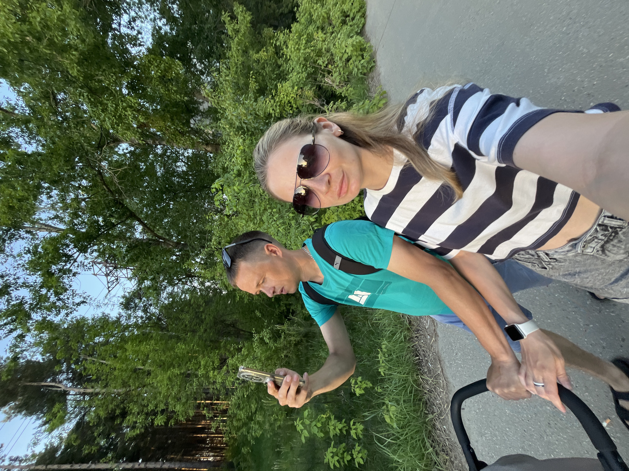 A woman and a man are outside on what appears to be a sidewalk or paved pathway. The woman is wearing sunglasses and a striped shirt. The man is wearing a teal shirt and has sunglasses perched on his head. He is holding a phone in his hand. Both are pushing a stroller. They are near a grassy area and trees. The location is Chaikovsky, Russia.