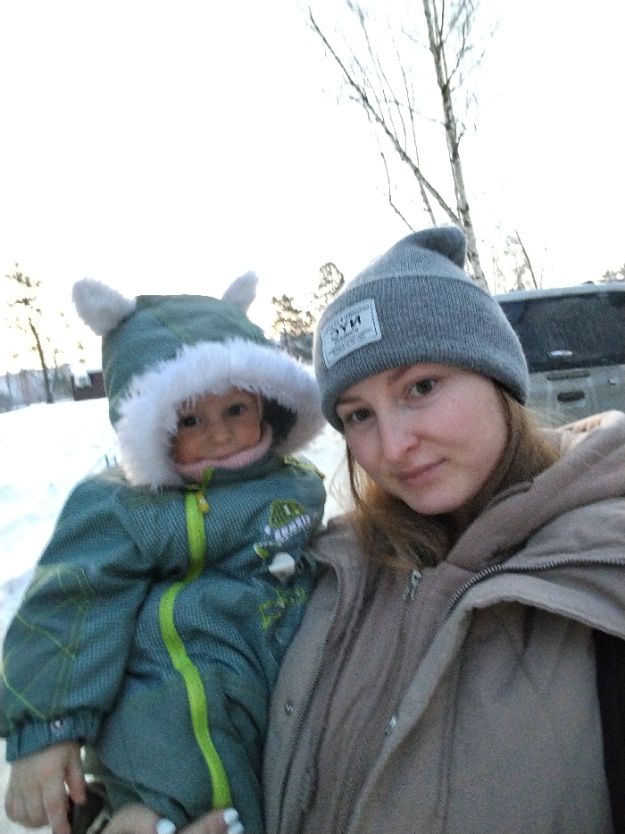 A woman in a gray beanie and tan jacket holds a child bundled in a green snowsuit with a furry hood. The child's suit features a dinosaur appliqué and neon green accents. They are standing outdoors in a snowy landscape with bare trees and a vehicle visible in the background. The lighting suggests either dawn or dusk. Both are looking at the camera.