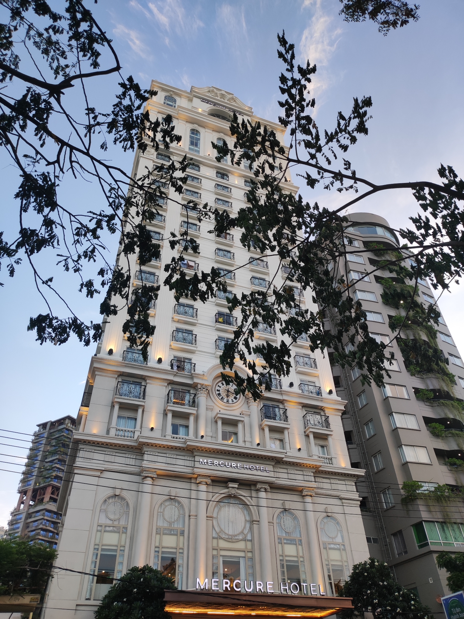 This media file depicts the Mercure Hotel building in Nha Trang, Vietnam, viewed from a low angle and framed by tree branches. The sky is visible with some scattered clouds, suggesting it is daytime, possibly late afternoon or early morning given the lighting. The main subject is the white, ornate Mercure Hotel, characterized by multiple floors with balconies featuring decorative railings. A large clock is prominently displayed on the facade of the building. Adjacent to the white hotel is a taller, curved building with a modern design, featuring numerous windows and balconies with greenery, indicating a potential integration of nature with urban architecture. The scene is an urban setting, likely a street in Nha Trang, with power lines visible in the foreground, adding to the realism of the location. There are no people clearly visible in the frame, and the focus is on the architecture and the surrounding environment. The overall mood is serene and observational, capturing the grandeur of the hotel against the backdrop of the sky and neighboring structures. The text "MERCURE HOTEL" is visible multiple times on the building, clearly identifying the establishment.
