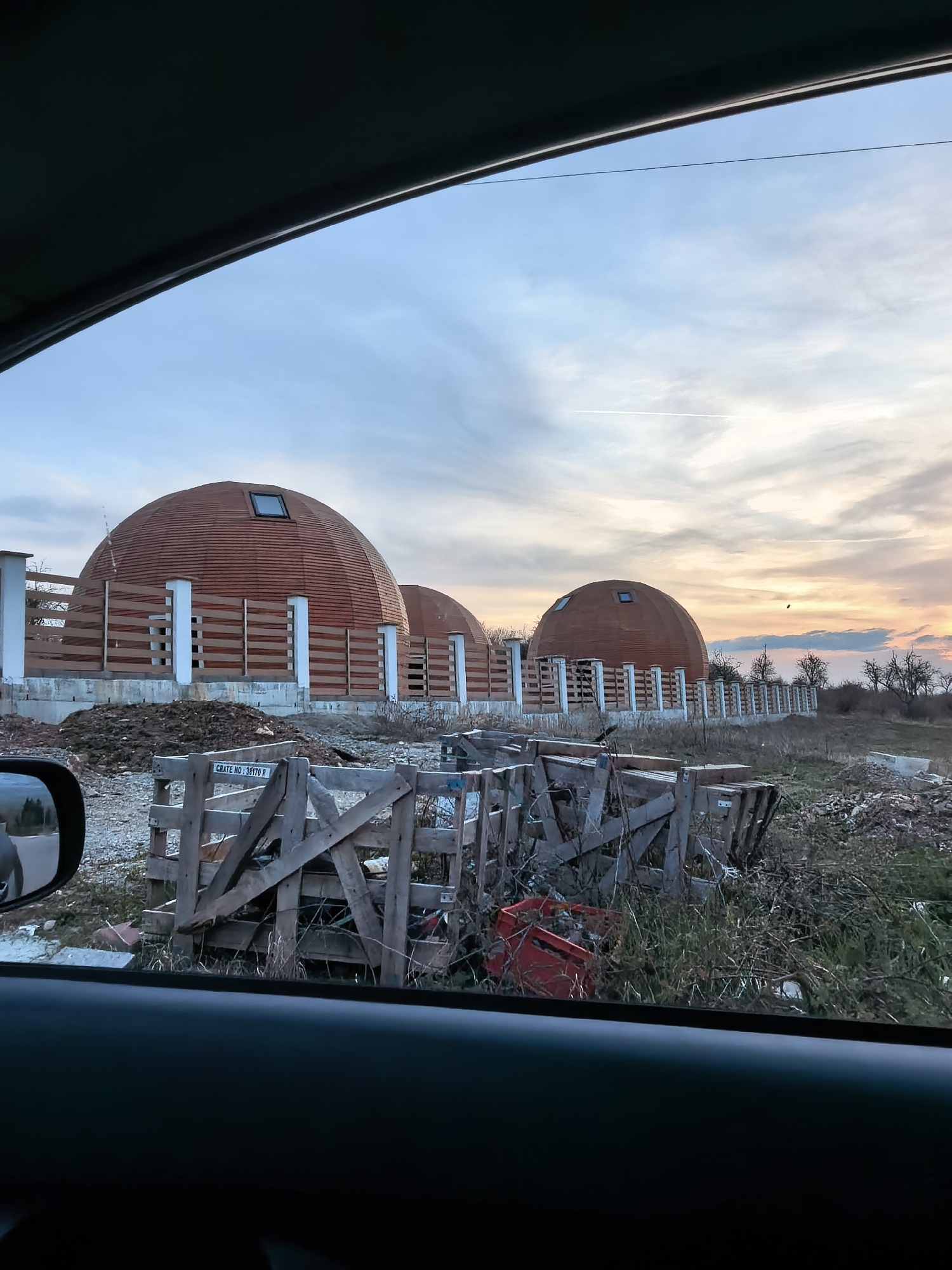 View from inside a car, looking out onto three dome-shaped buildings with brown exterior cladding. Each building features a white post and beam fence structure. The foreground contains wooden crates and assorted debris. The sky is overcast with hues of blue and orange from the setting sun. The location appears to be a rural or suburban area.
