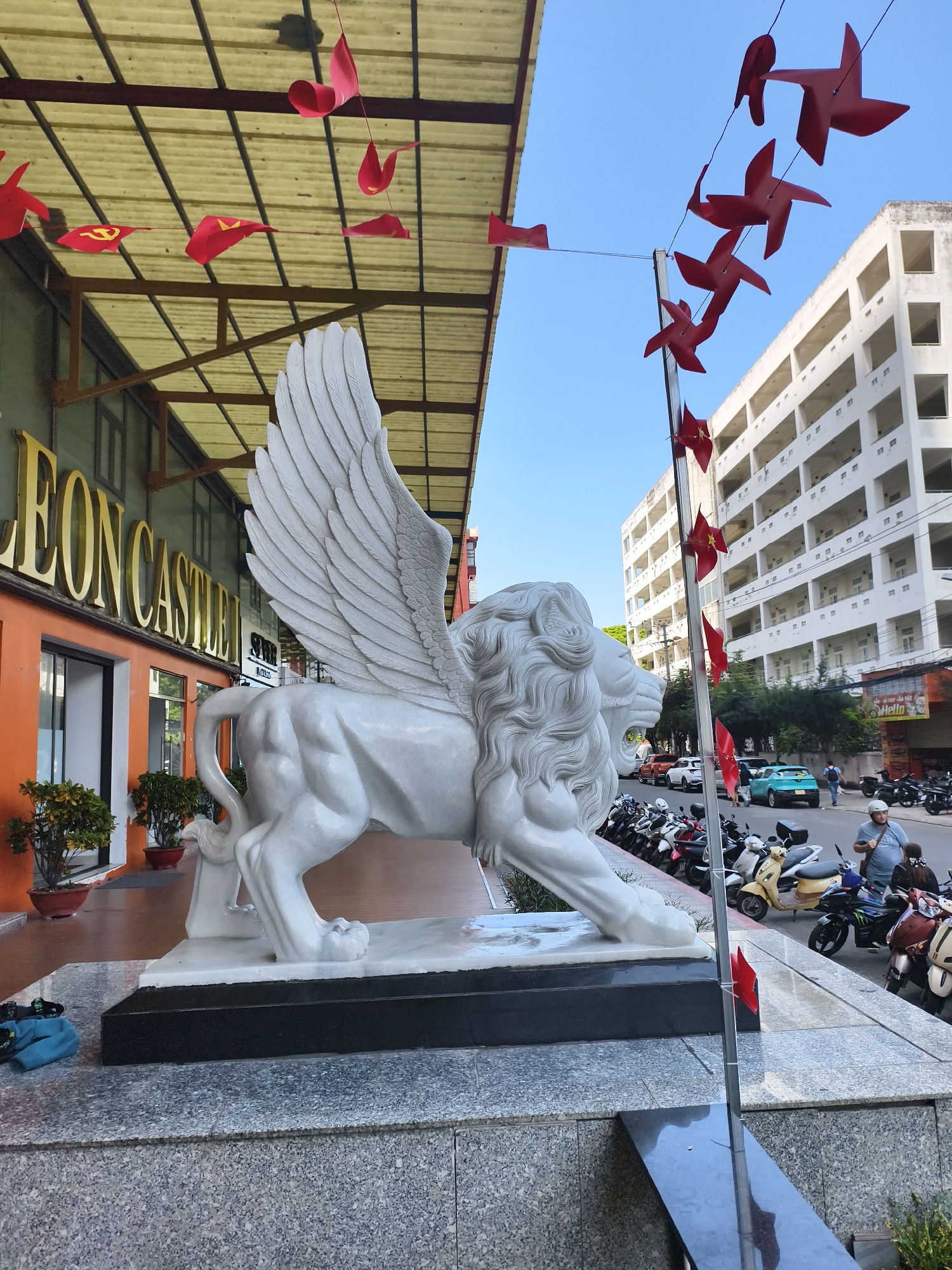 The image captures a street scene in Bac Nha Trang Ward, Vietnam. Dominating the foreground is a large, white marble statue of a winged lion, positioned in front of a building with the words "LEON CASTLE" in gold lettering. Red flags are strung overhead, adorning both the building and a nearby pole. The street is lined with parked motorcycles, and pedestrians are visible in the background, including a man wearing a helmet and riding a scooter. A multi-story white building is also visible across the street. The sky is clear and blue.