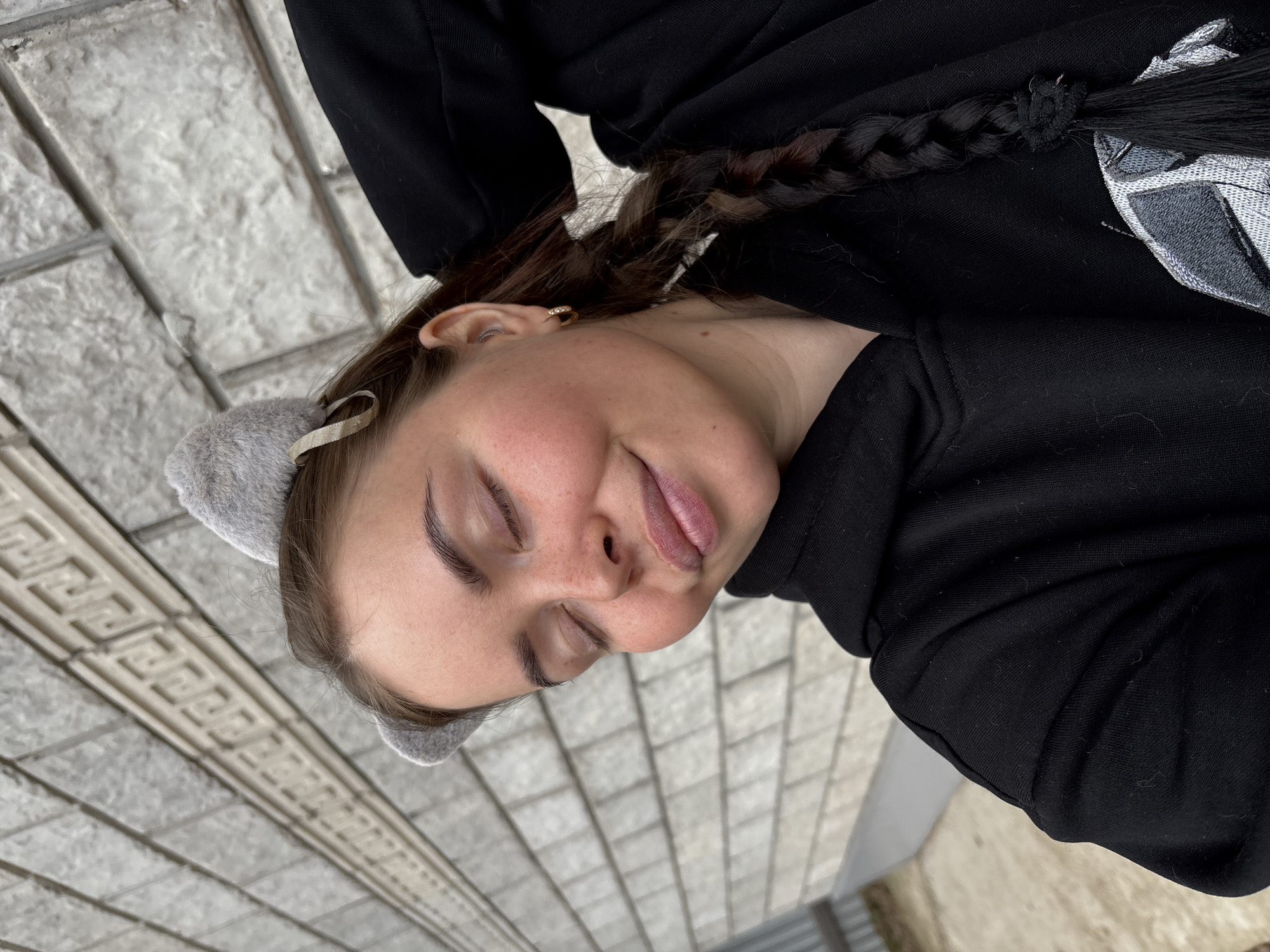 A woman with a braid is lying down on a brick surface in Русский Вожой, Russia. She is wearing a black top and fuzzy cat ears. She has her eyes closed.