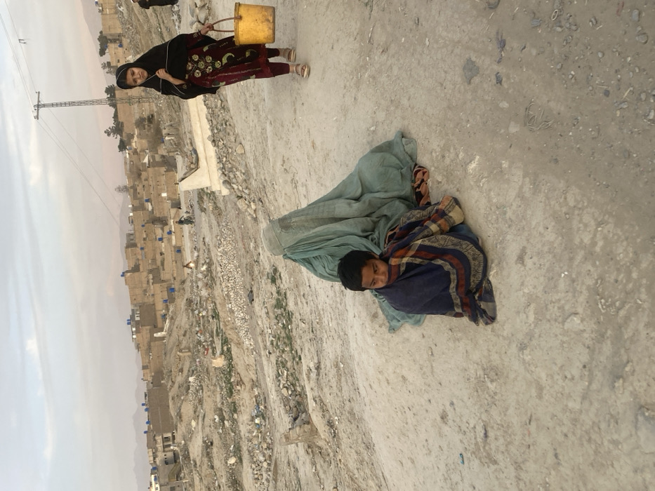This image depicts a dusty, arid landscape within Quetta City Tehsil, Pakistan, characterized by a settlement in the background and a grave site in the midground.
In the foreground, a young child, likely male, with short dark hair and a solemn expression, sits on the unpaved, light brown ground. He is wrapped in a dark blue blanket or shawl with red and beige stripes, covering his body and drawn up around his lower face. Directly behind him and slightly to the right, an adult figure is seated on the ground, completely enveloped in a light greenish-blue burqa, obscuring their identity.
To the right, on an unpaved path running diagonally across the frame, a young girl walks from right to left. She wears a dark head covering, a dark maroon tunic with green embroidered patterns, and light-colored sandals. In her right hand, she carries a yellow plastic bucket with a handle. Her gaze is directed downwards and slightly to her left.
The midground features undulating, barren terrain littered with small rocks, sparse dry vegetation, and scattered debris. Several unkempt grave markers, appearing to be white concrete or stone structures, are visible throughout this area, some partially obscured or damaged. Further back on the right, a tall metal electricity pylon with overhead power lines stands against the sky.
In the background, a dense cluster of low-rise, light-colored mud or brick buildings forms a residential area, typical of the region. Many rooftops display blue plastic barrels or containers. Distant hills or mountains frame the horizon under a hazy or overcast sky. A figure, likely an adult male, is partially visible standing further back on the right side of the background. A lone individual on a bicycle is also visible traversing a path within the background settlement.