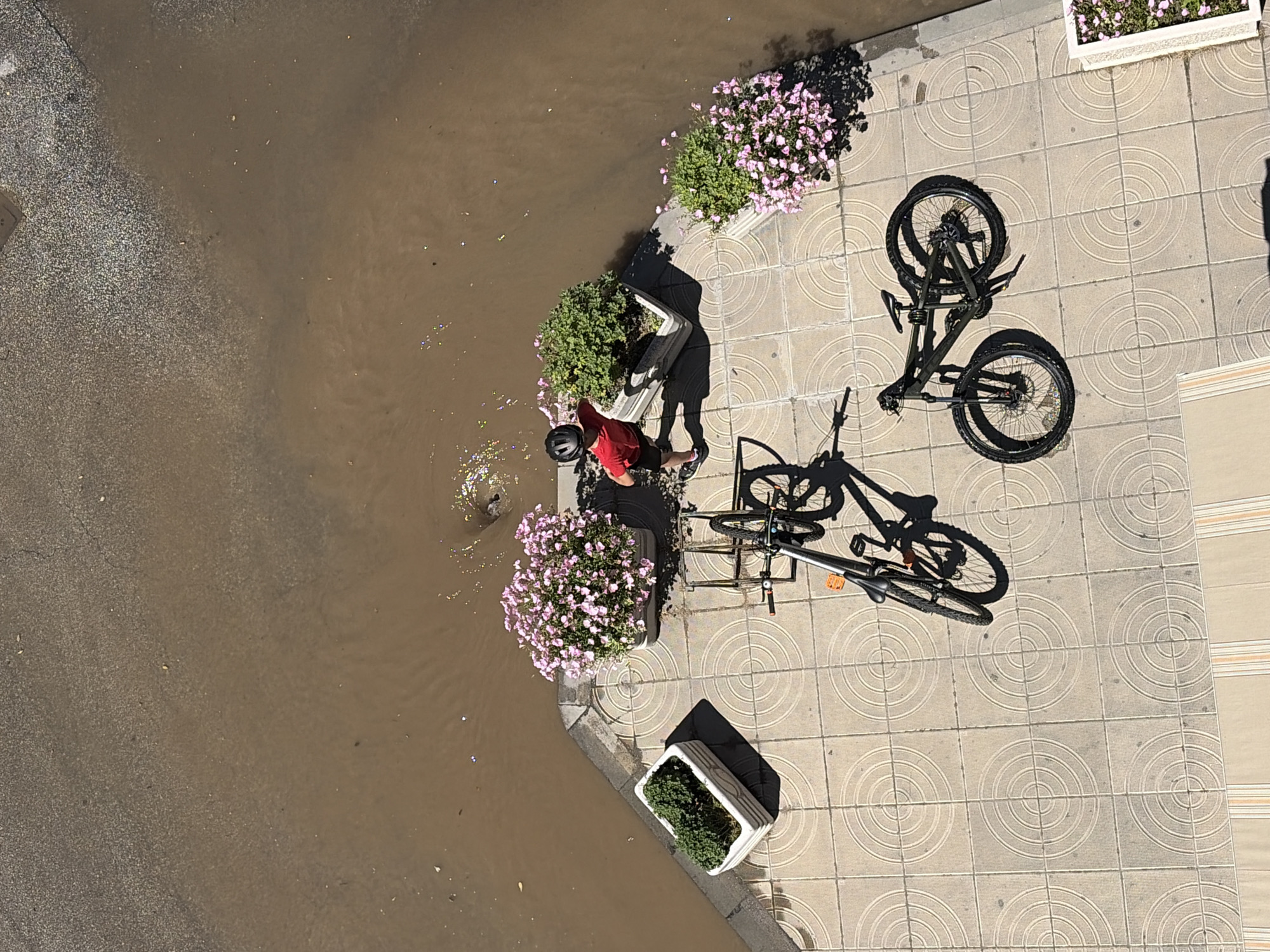 An aerial photograph shows a flooded street in an urban area. The water, muddy and brown, covers a significant portion of the left side of the frame. Along the edge of the street, a paved sidewalk is visible, featuring a repeating circular pattern on the tiles.
Along the sidewalk, there are several planters containing flowers and greenery. A person wearing a red shirt, black shorts, and a helmet is standing near a planter, seemingly interacting with the water. Two bicycles are parked on the sidewalk, one lying on its side. The location is unknown.