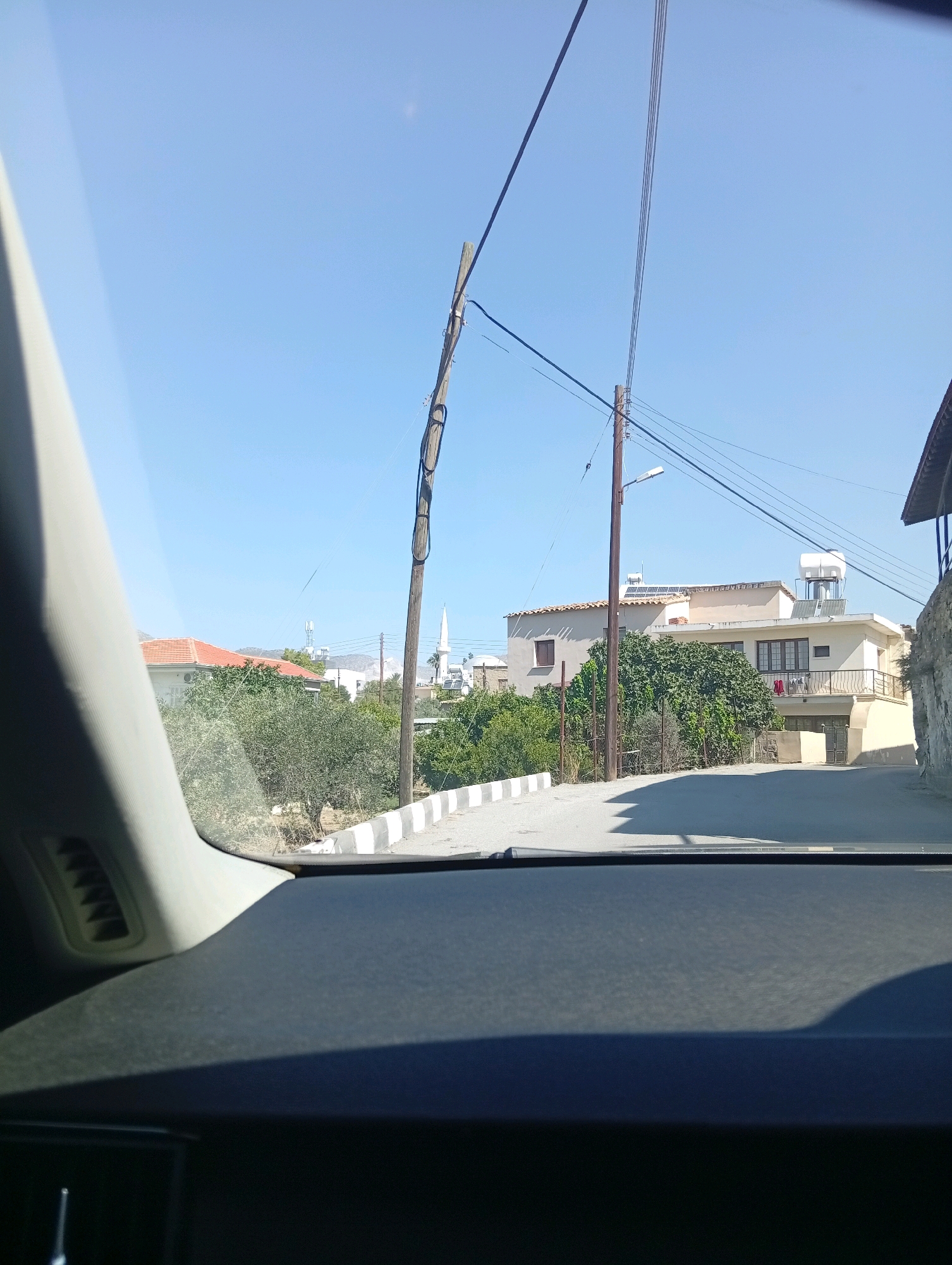 The image captures a view from inside a vehicle looking out onto a street in Değirmenlik, Northern Cyprus. The scene is dominated by a bright, clear blue sky. A road with white and black striped curbs runs diagonally from the lower right to the upper left of the frame. On the left side of the road, a car's interior dashboard is partially visible, along with a portion of the windshield.
Across the road, several buildings are present. A house with a red tiled roof is visible on the left, surrounded by dense green foliage. Further along the road, a cream-colored building with multiple windows and balconies is prominent, with solar panels on its roof and a white water tank. In the background, a tall, slender minaret is visible, suggesting the presence of a mosque or religious building.
Several utility poles with numerous wires are scattered throughout the scene, contributing to the rural or suburban feel of the location. The overall impression is one of a sunny day in a residential area, likely in a Mediterranean or similar climate.
