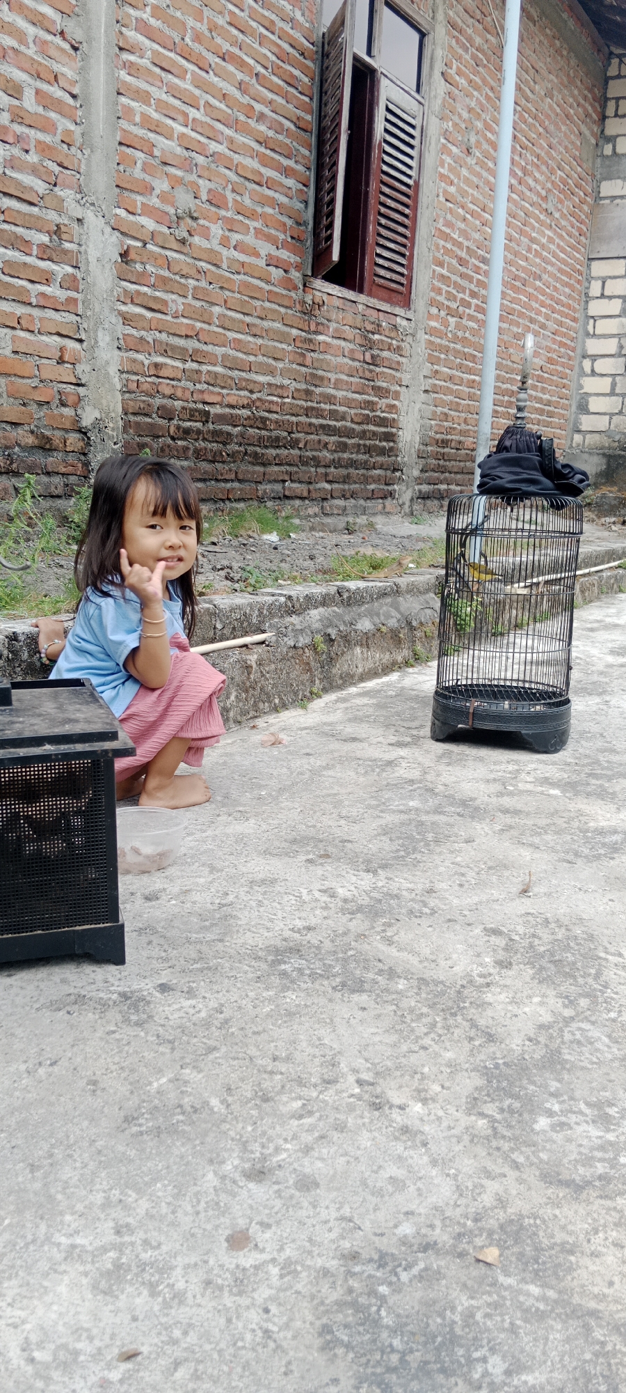 A young girl is sitting on a concrete ledge in front of a brick wall, with a birdcage to her right. She is wearing a blue t-shirt and a pink skirt. Her hair is dark and falls to her shoulders. She is looking at the camera and holding up her hand in a gesture. In the birdcage, there is a small yellow bird. To the left of the girl, there is a black cage-like object. The environment appears to be outdoors, possibly in a residential area. The brick wall is old and weathered, with some patches of exposed concrete. The ground is a rough concrete surface. There is a thin, light-colored pole extending vertically behind the birdcage, likely a utility pole. The image is taken at eye level, suggesting the photographer is standing or kneeling. The lighting is overcast, creating a soft and diffused light across the scene. It is difficult to ascertain the exact location within Pelang, Indonesia, without further context, but the architectural style of the brick wall and the presence of what appears to be a bird, commonly kept as pets or for singing competitions in some Southeast Asian cultures, could suggest a neighborhood setting.