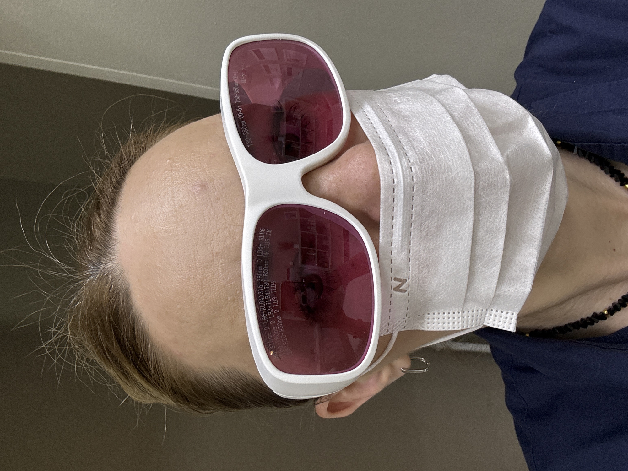 The image shows a person wearing a white surgical mask and white-framed glasses with dark pink lenses, looking down towards the camera. The glasses have text printed on them indicating laser protection. The person is wearing a dark blue garment and a black beaded necklace with gold accents is visible. The background is a plain light gray. The context of the image does not suggest any connection to the city of Murino, Russia.