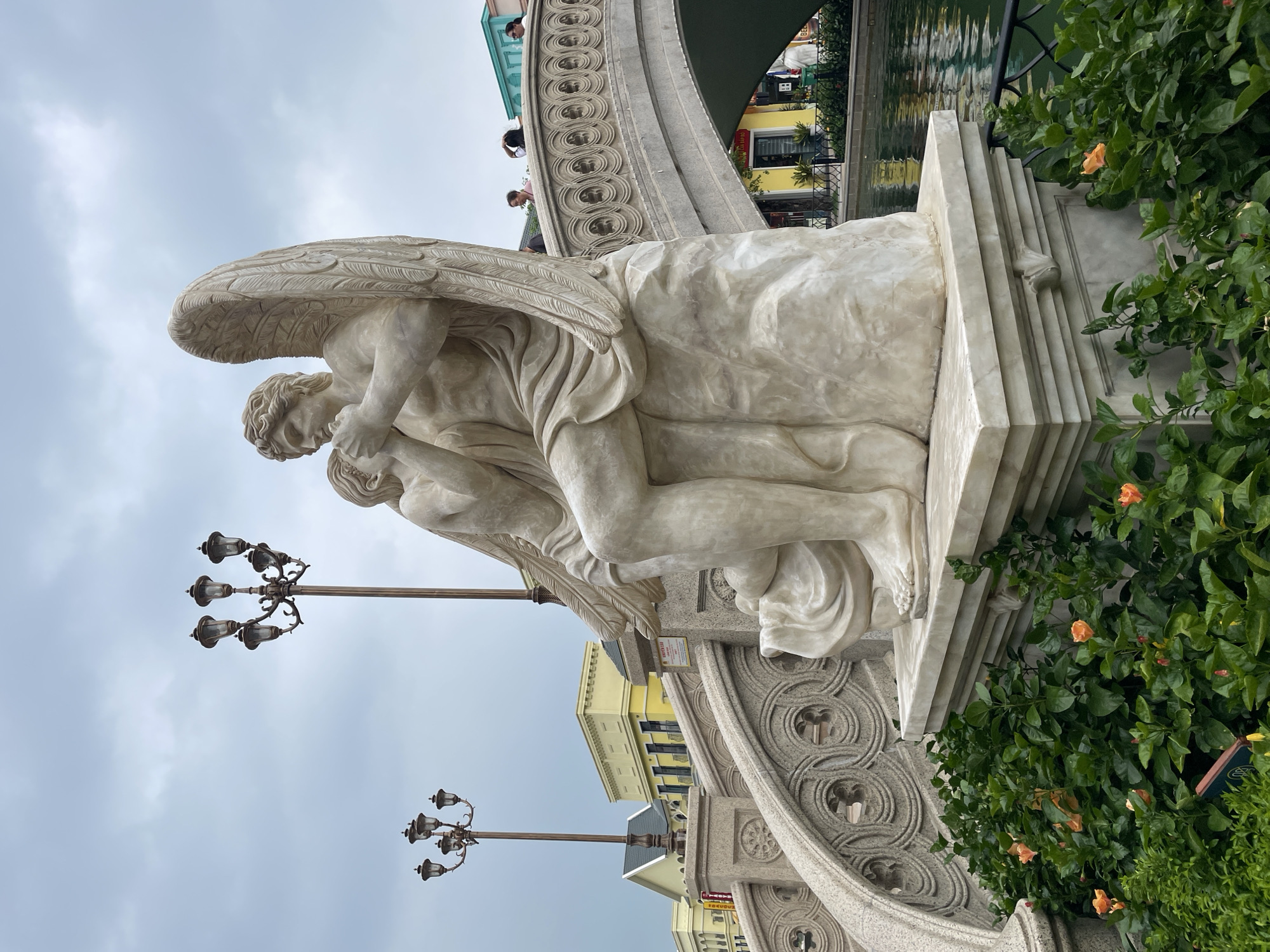 The image depicts a marble sculpture of two figures, likely an angel and a child, situated in front of a body of water. The angel, with large wings, is seated and appears to be embracing the child. The sculpture rests on a marble pedestal adorned with classical motifs. Behind the sculpture, a bridge with intricate stonework arches over the water. On the bridge and in the background, several people are visible, as well as ornate lampposts and buildings with yellow and green facades. Lush green bushes with orange flowers surround the base of the sculpture and the water's edge. The sky is overcast. This scene is located at the Hoi An Impression Theme Park in Nghĩa Trụ Commune, Vietnam, a place designed to evoke a sense of historical European charm.