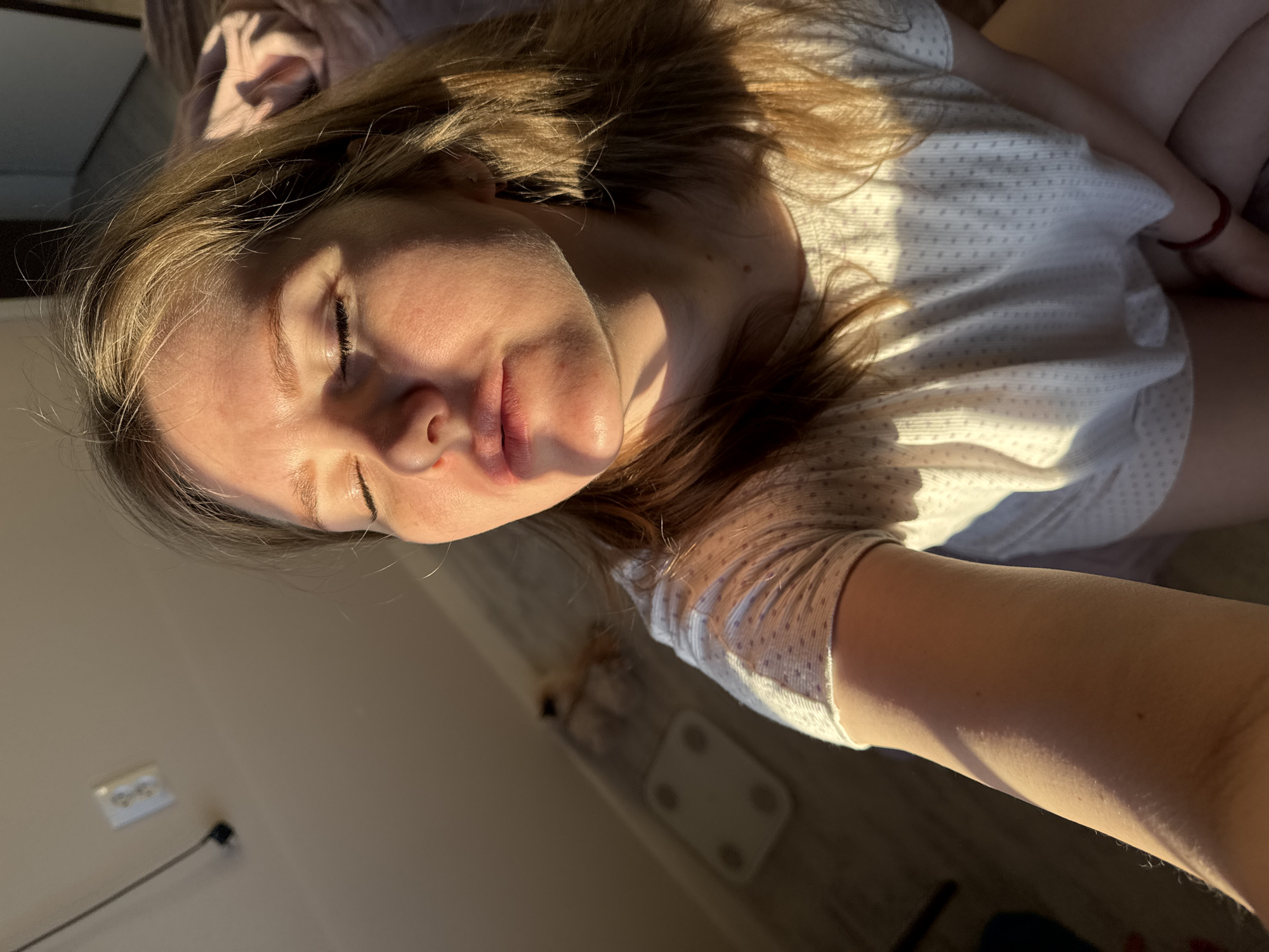 A young woman with long, light brown hair is shown resting indoors, possibly in a bedroom or living room, illuminated by direct, golden sunlight. She is wearing a light-colored, patterned t-shirt, and her eyes are closed or partially closed, conveying a sense of relaxation and peace. The strong, low-angle light indicates either early morning or late afternoon.
The setting includes a wooden or laminate floor, a white power outlet with a plugged-in cord on the wall, and what appears to be a bathroom scale on the floor. A soft, textured fabric, possibly a blanket or pillow, is visible behind her head. The image is captured from an overhead, slightly tilted perspective. The scene is calm and serene, highlighted by the warm natural light. There is no visible text in the image. The location context provided is Gornyi (Dvurechenskoe S/P), Russia.