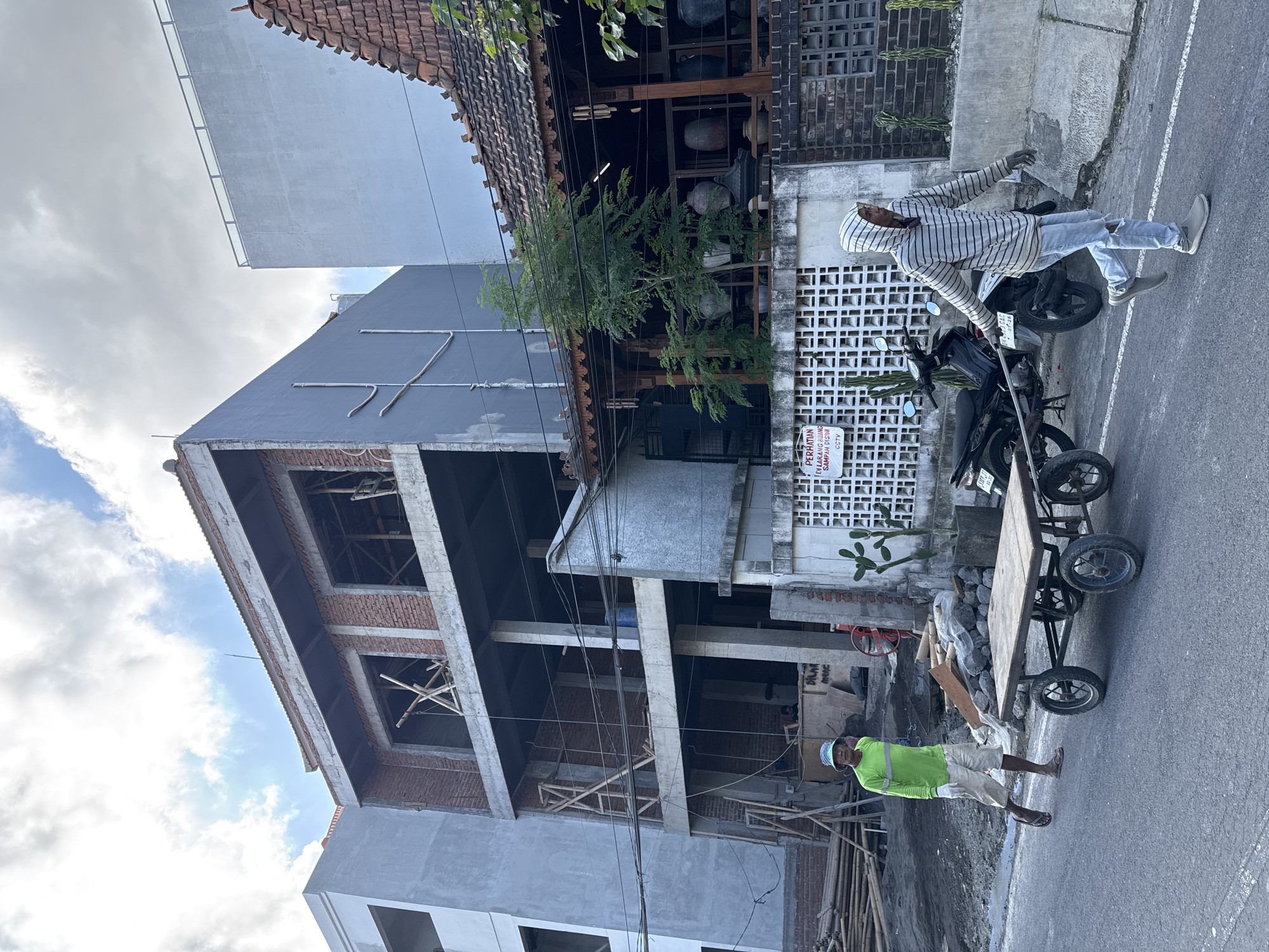 In North Kuta, Indonesia, on a street with a partly cloudy sky, a construction site is active. A multi-story building under construction, made of brick and concrete, dominates the background. Adjacent to it, a more traditional structure with a tiled roof and wooden decorative elements displays pottery.
In the foreground, two men are engaged in labor. One man, wearing a bright green long-sleeved shirt, beige shorts, white gloves, and a blue hat, walks past a handcart with wooden planks on its bed. The handcart has four wheels. The second man, wearing a striped hooded jacket, ripped light-blue jeans, and gray sneakers, is pulling the handcart from behind.
A black motorcycle is parked behind the handcart. A white wall with a decorative lattice pattern is visible, bearing a sign with text in Indonesian that reads "PERHATIAN DI LARANG BUANG SAMPAH DISINI CCTV" (Attention: Do not litter here. CCTV). Several cacti are planted in a small square planter near the wall. The street is paved with asphalt, and building materials like bamboo poles and sand are piled up on the left side of the image.